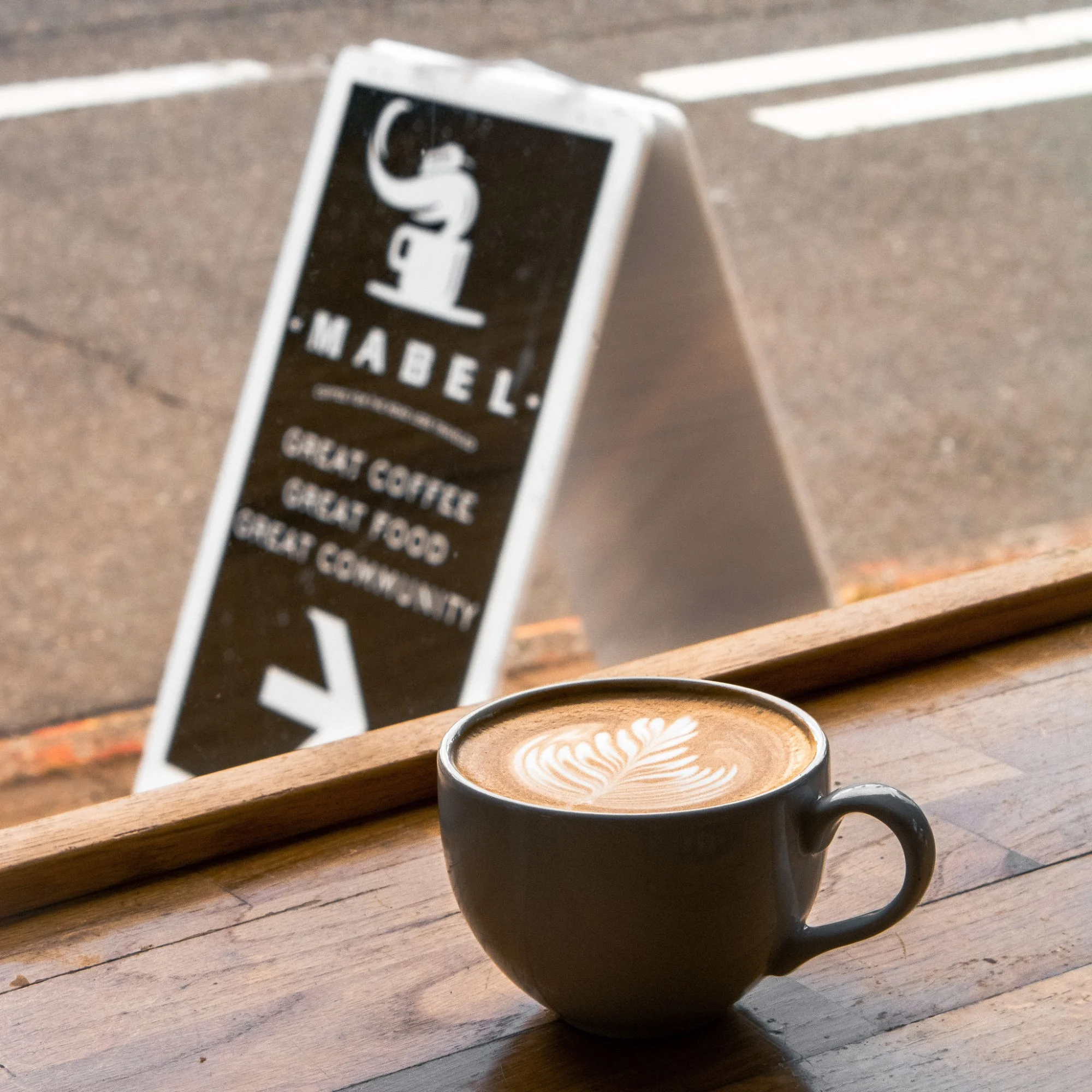 A cup of coffee with latte art on a wooden table near a window, with a sidewalk and a street sign in the background.