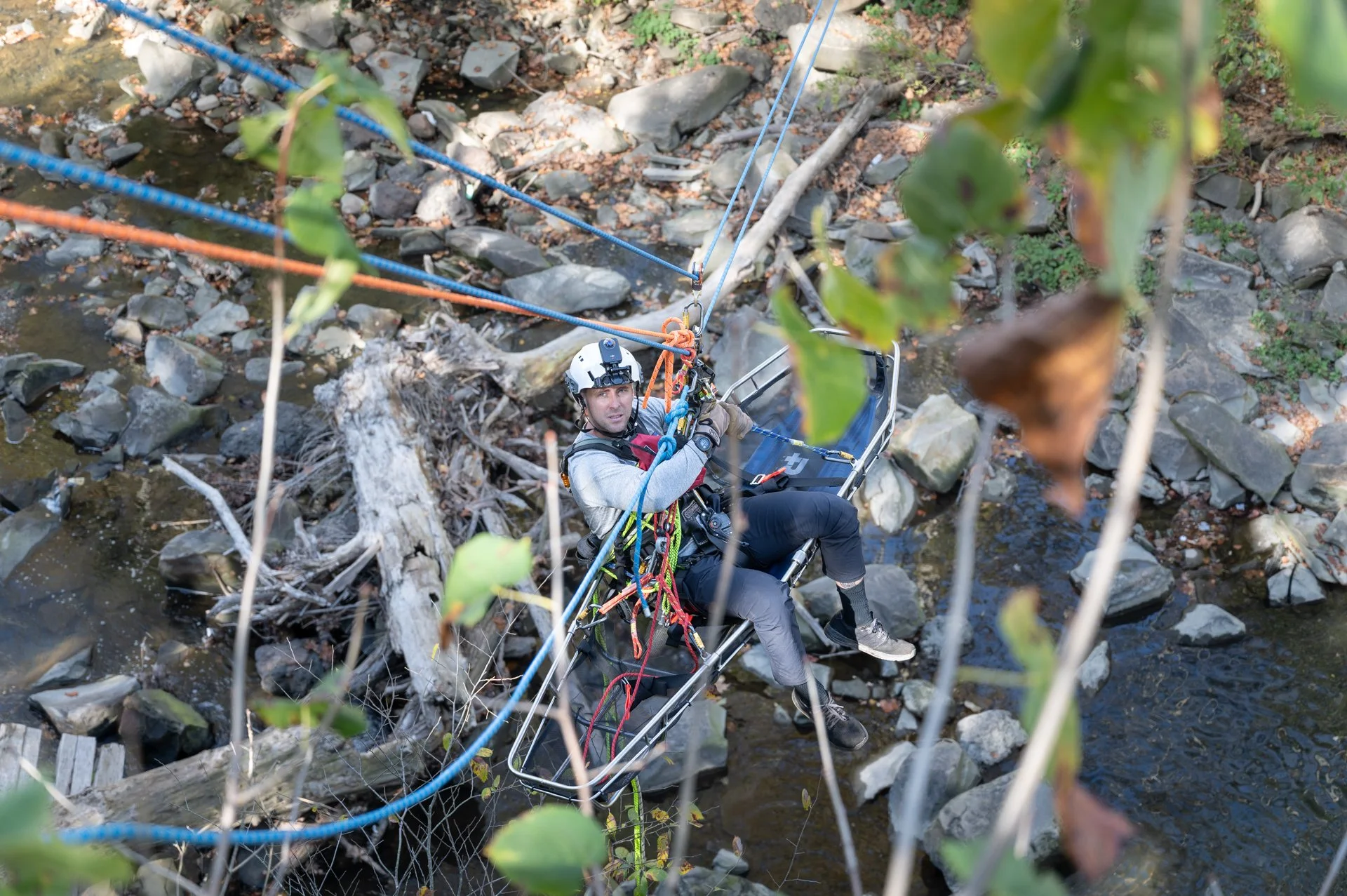 A man with climbing gear sitting on a rescue stretcher suspended over a rocky river, surrounded by trees and leaves.