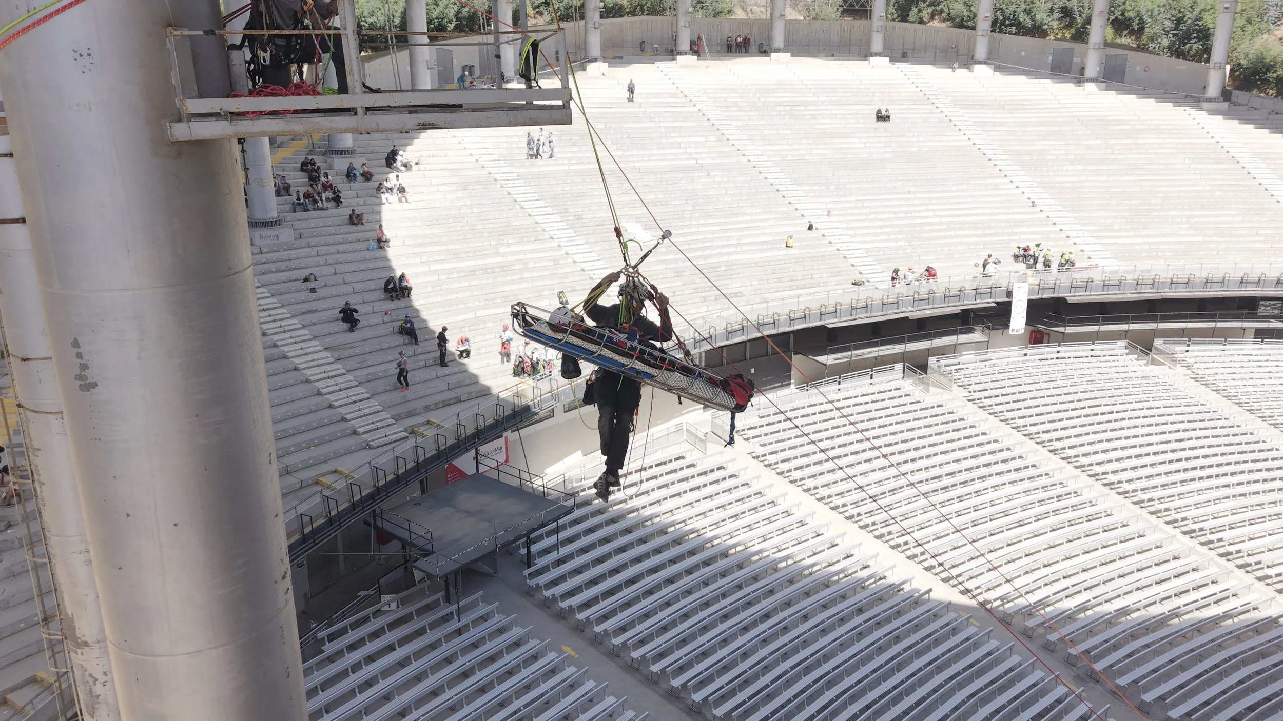 Construction worker hanging from a platform suspended above stadium seating during construction at a large outdoor sports arena.