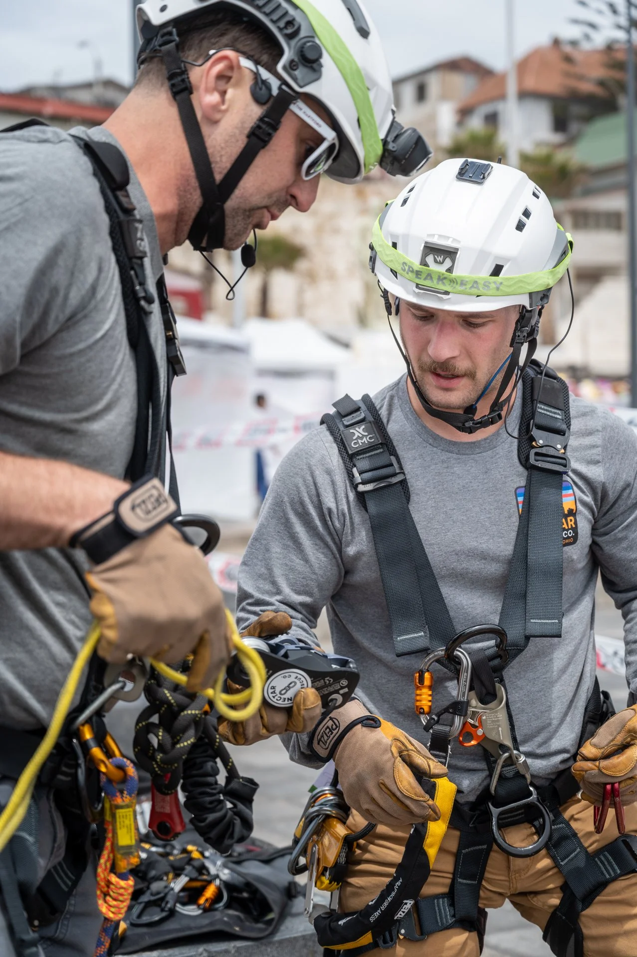 Two rescue workers wearing helmets, harnesses, and gloves discuss equipment outdoors.