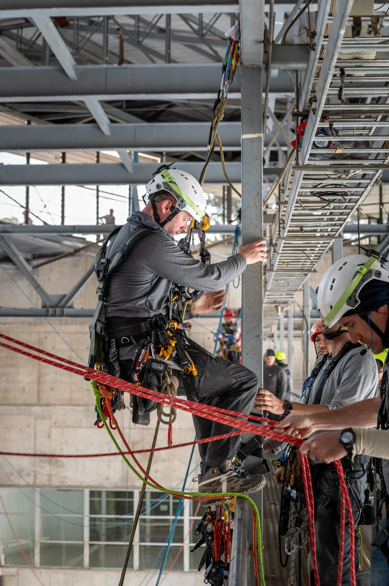 A worker in safety gear, helmet, and harness preparing to rappel from a scaffold at a construction site, with other workers and equipment visible in the background.