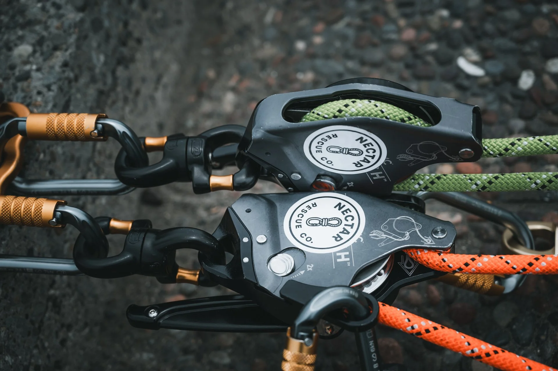 Close-up view of climbing equipment, showing two linked carabiners with orange and black textured ropes, attached to a black device labeled 'NEGTAH RESCUE CO.,' on a rocky surface.