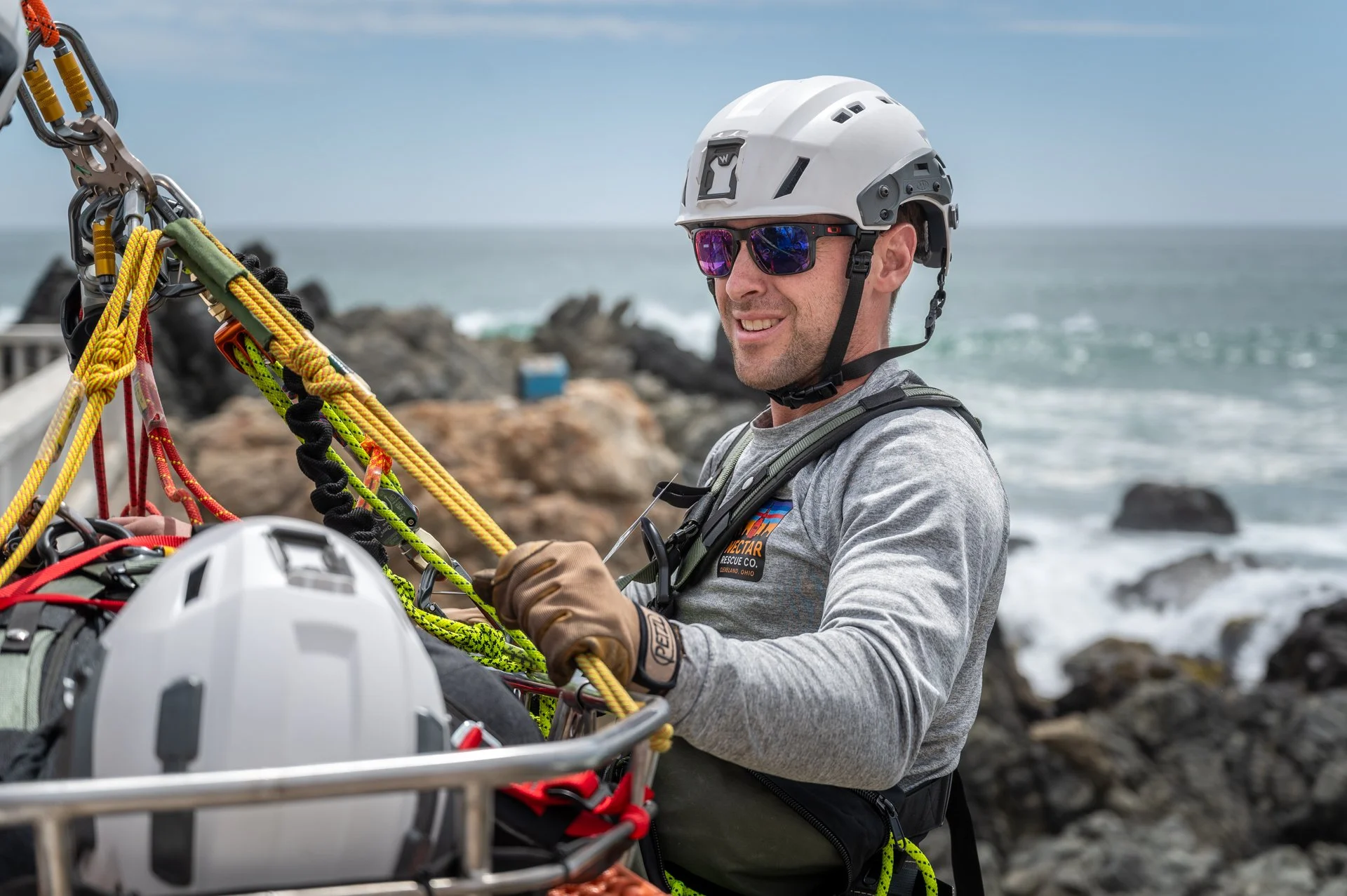 Rescue worker in gray shirt, white helmet, and sunglasses handles rescue equipment outdoors near the ocean with rocks in the background.
