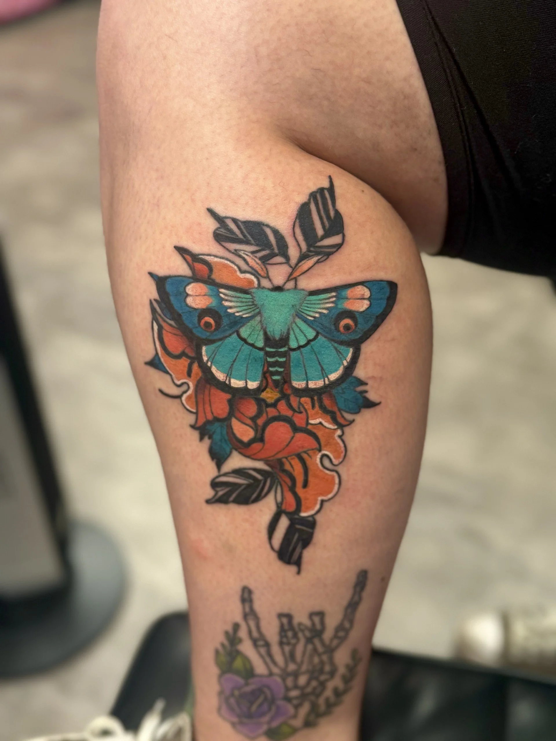 Tattoo of a colorful butterfly on a person's calf with black, orange, blue, and green wings, surrounded by floral and stem designs.