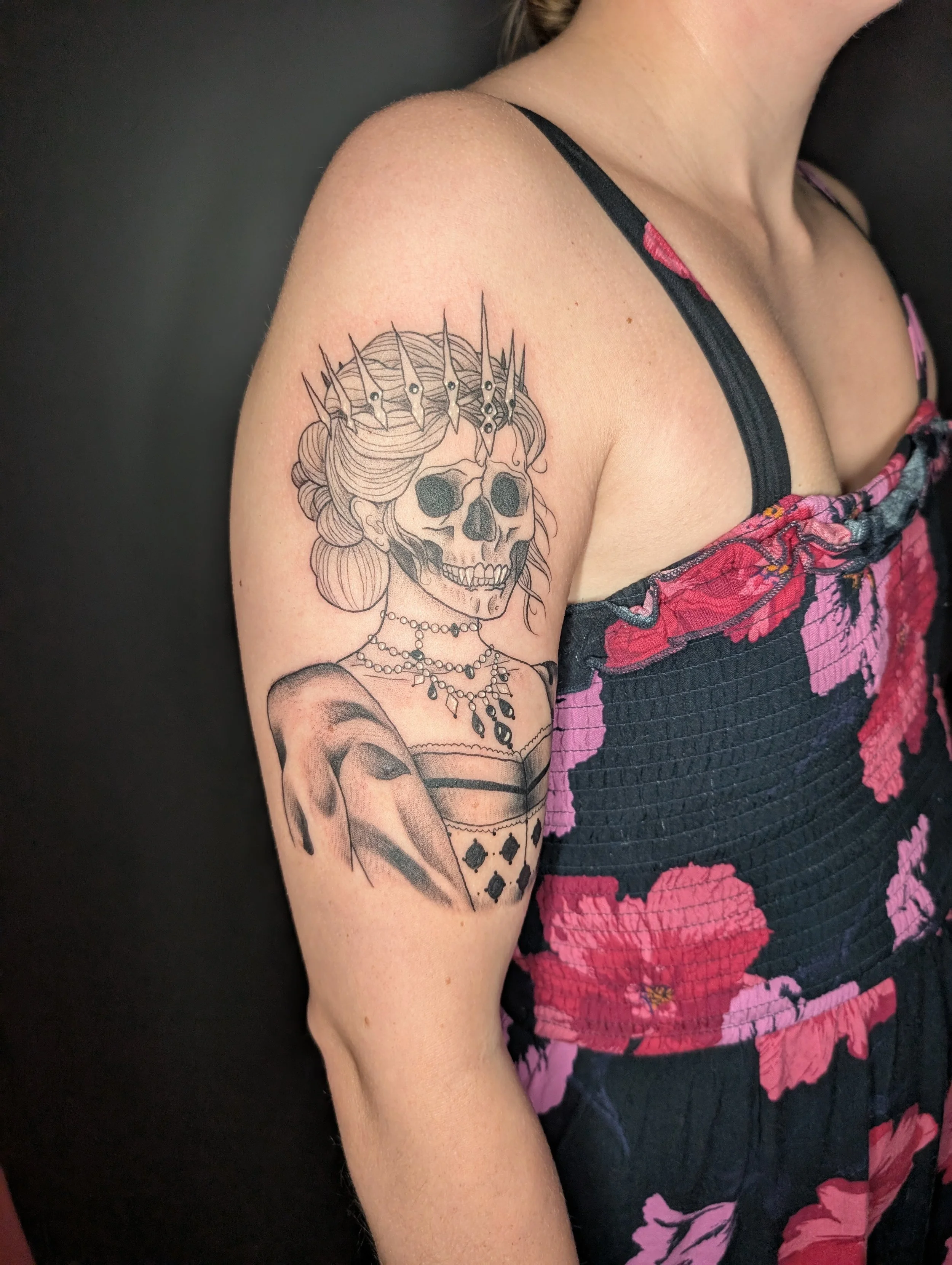 A woman with a tattoo of a skull wearing a crown of spikes and a necklace on her upper arm, wearing a sleeveless shirt with pink and black floral pattern.