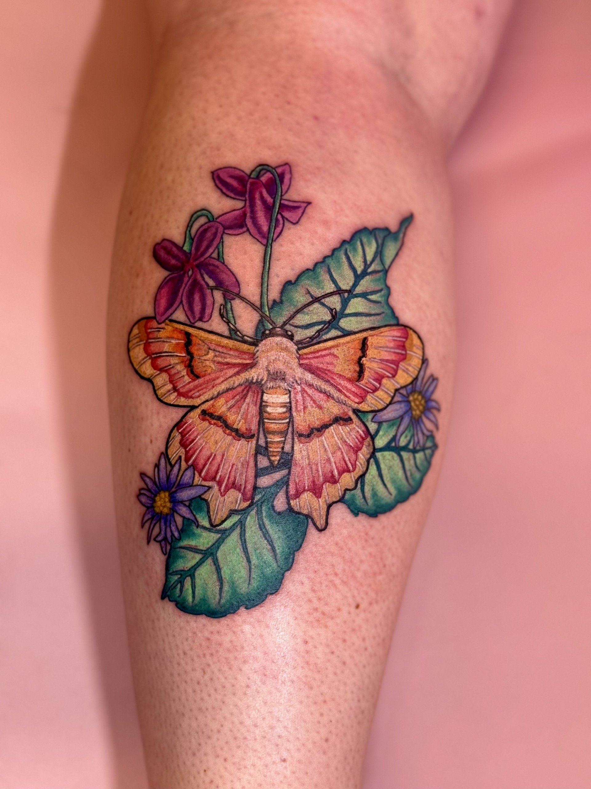 Colorful tattoo of a moth, purple flowers, and green leaves on a person's arm.