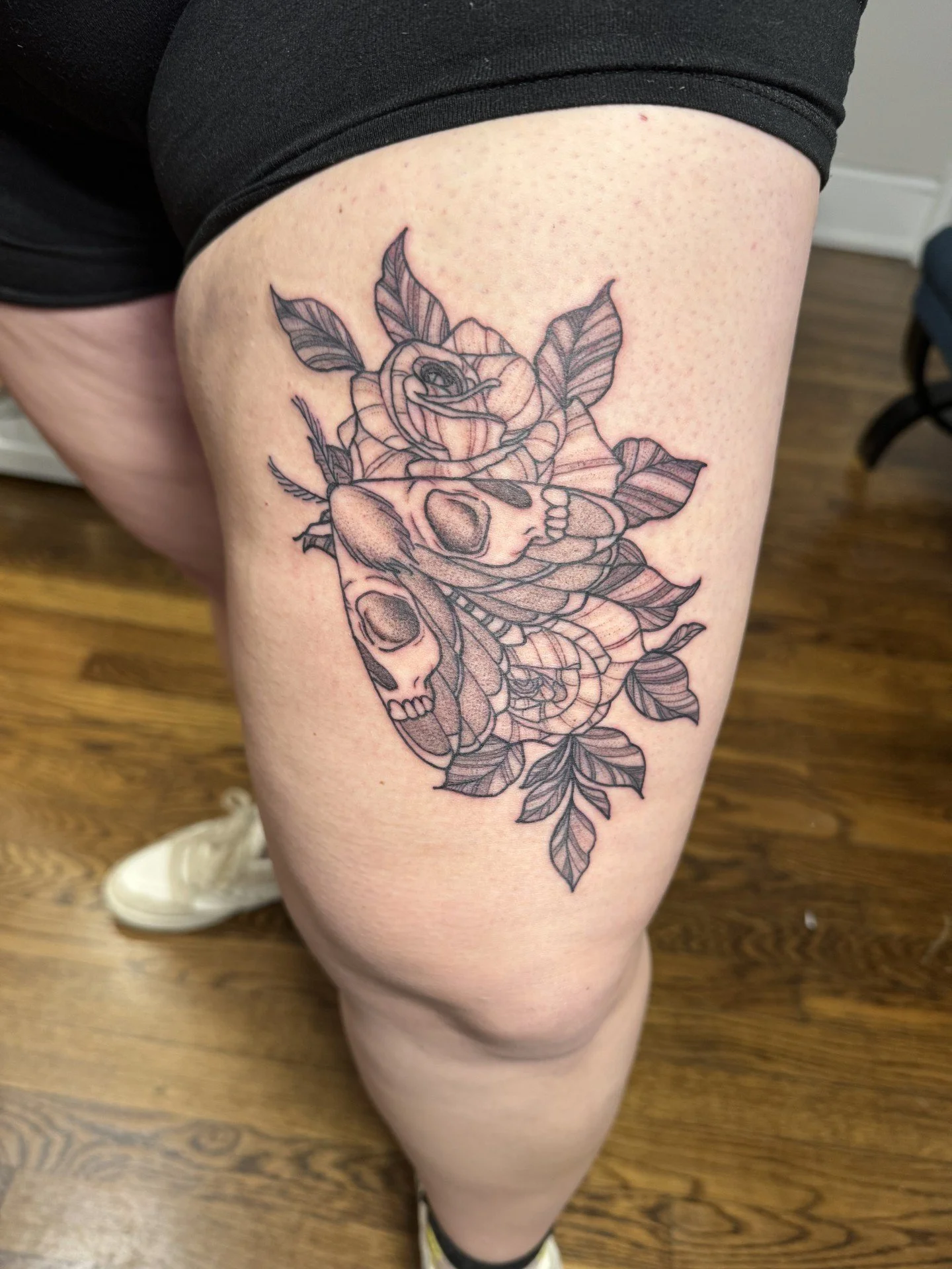Tattoo of a rose with skulls, leaves, and stems on a person's thigh.
