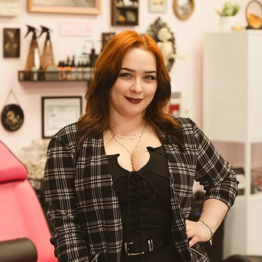 A woman with red hair and dark lipstick standing indoors, wearing a black top with a black and white checkered blazer, smiling slightly.