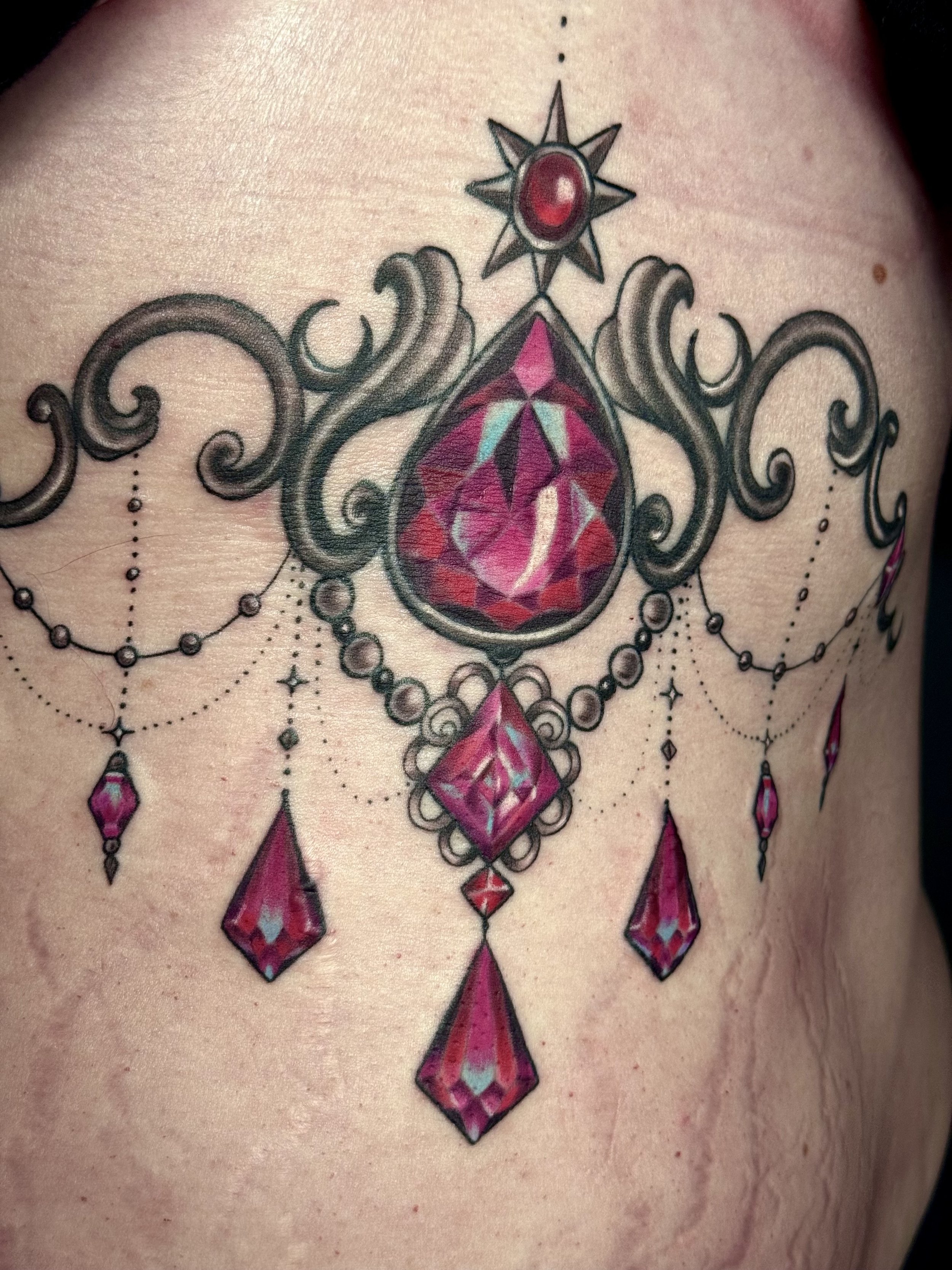 A colorful tattoo of a large red gemstone with ornate black and pink design elements, including dangling jewelry-like shapes, on skin.