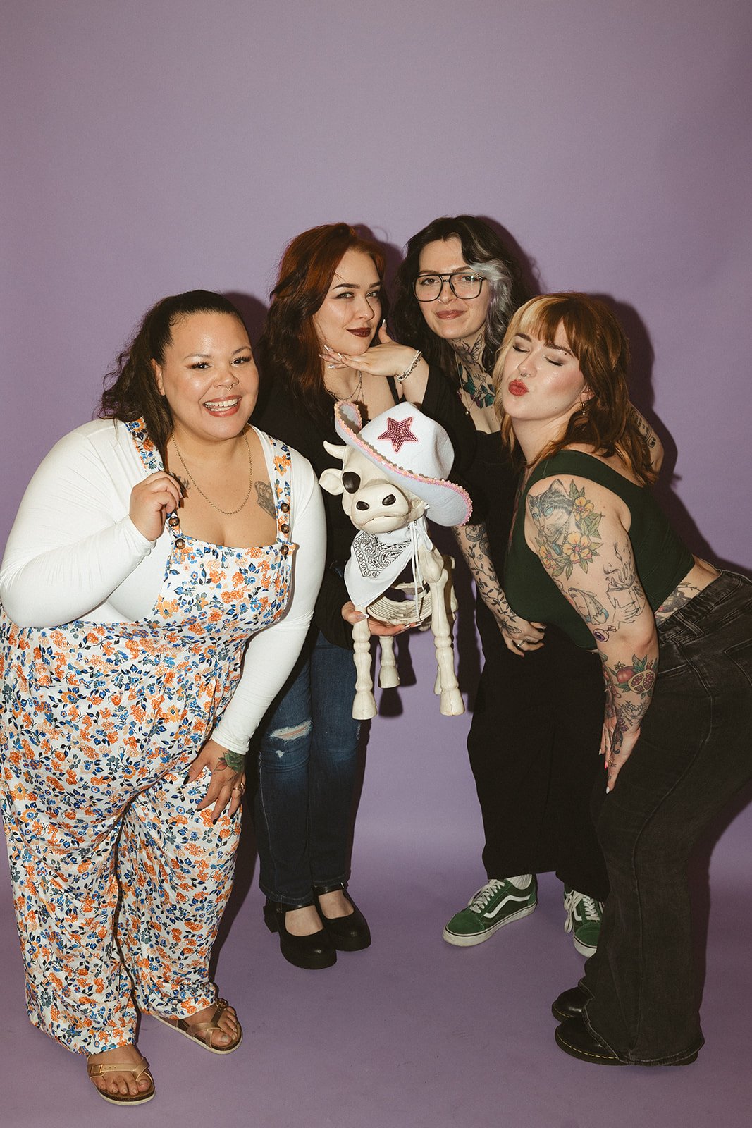 Four pretty women covered in tattoos posing with a skeleton cow dressed as a cowboy, against a purple background.