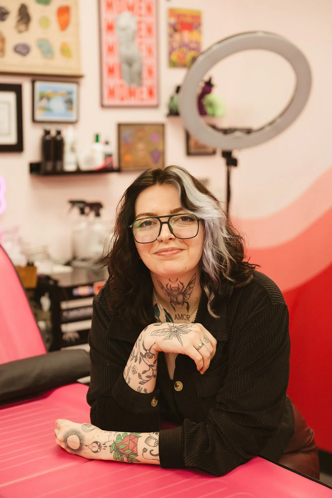 A woman with glasses and tattoos smiling at the camera in a tattoo studio with framed art on the wall and a ring light in the background.