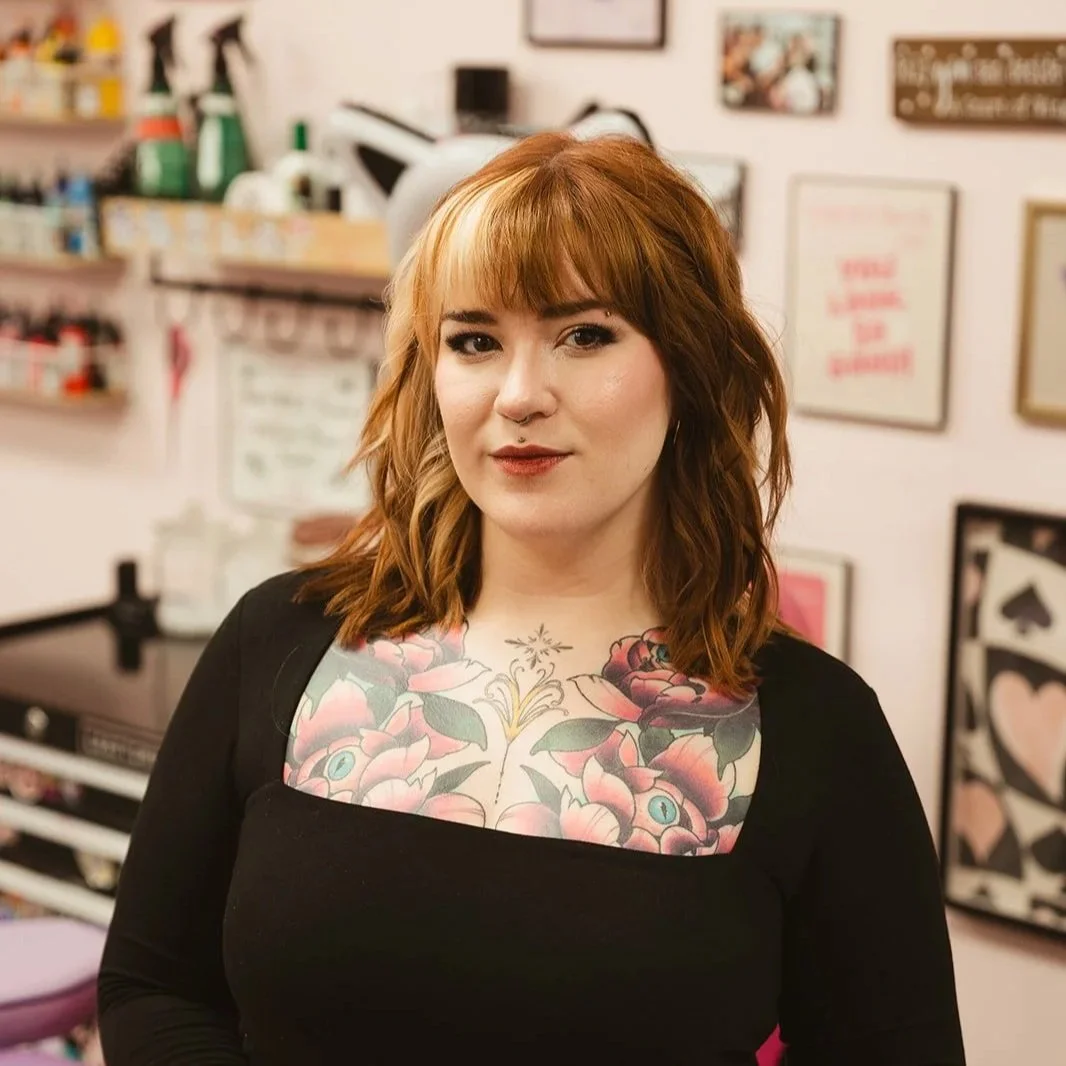 A woman with shoulder-length wavy reddish-brown hair and tattoos on her chest, wearing a black top, stands in a tattoo studio with framed artwork and supplies in the background.