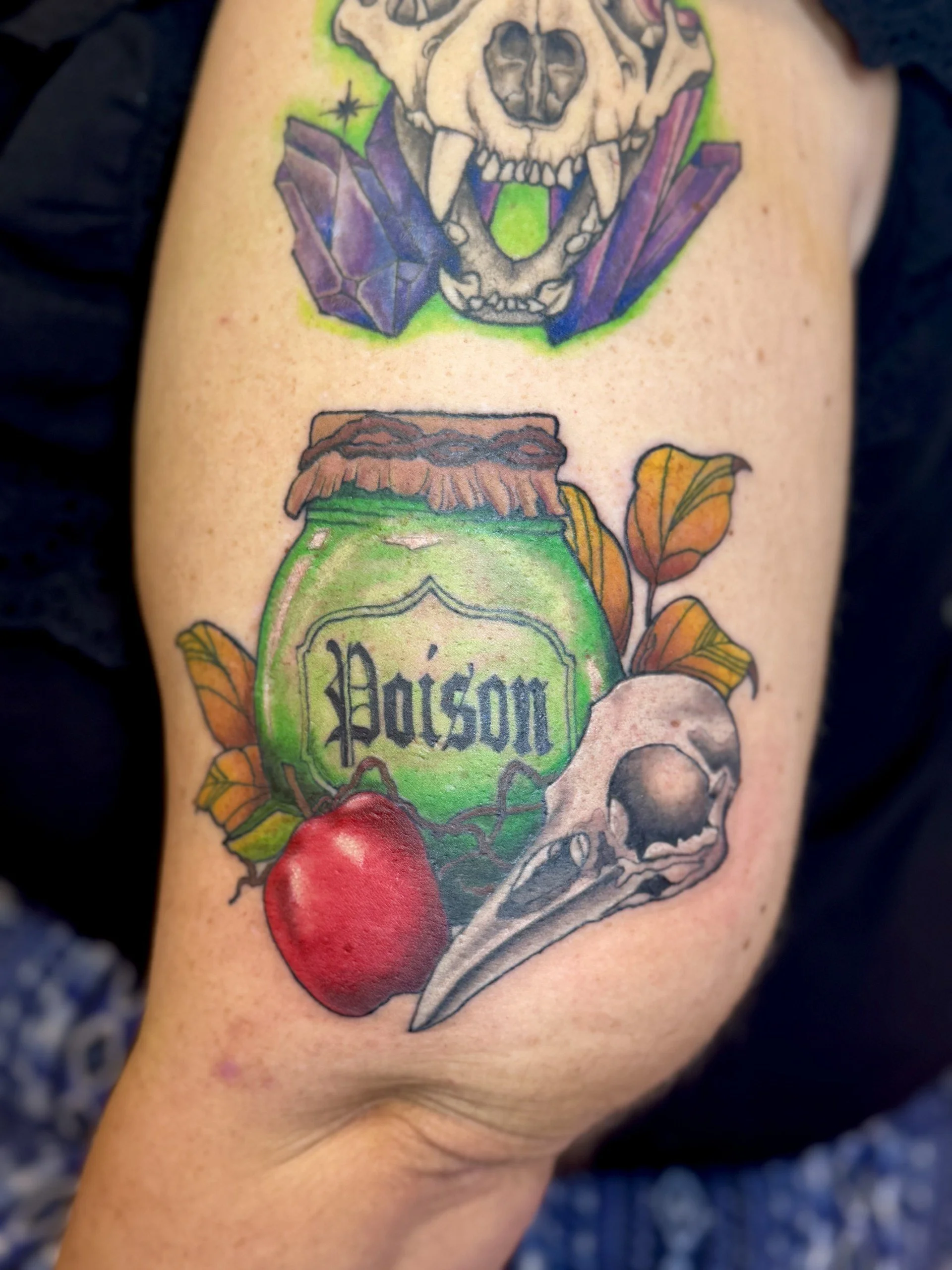 A tattoo on a person's upper arm featuring a skull with a dog face, purple crystals, a green label with the word 'Poison,' a red apple, leaves, and a dagger.