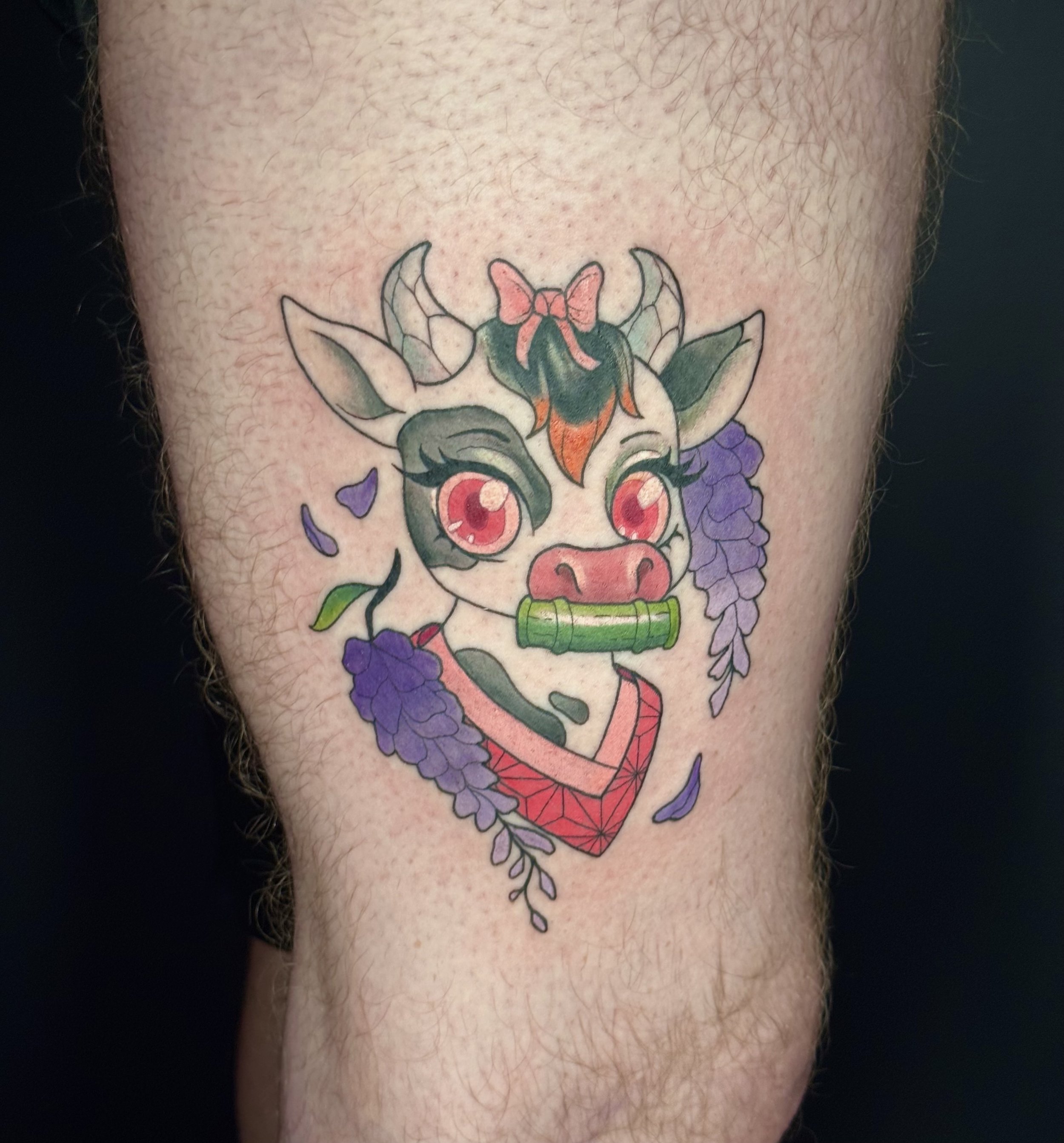 Colorful tattoo of a cartoonish style cow with a pink bow on her head, surrounded by purple flowers. It references an anime character