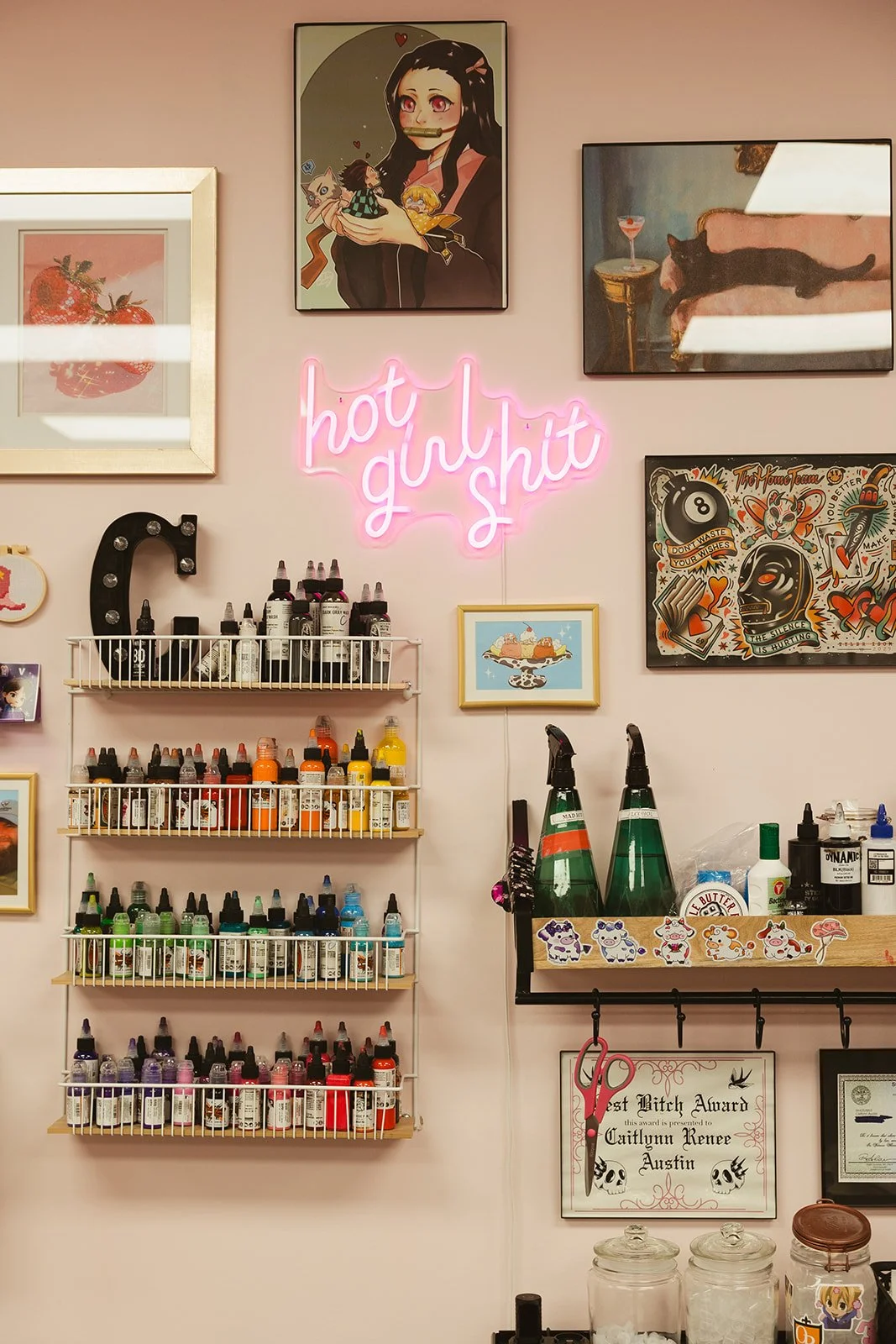 Decorative wall with artwork, a pink neon sign saying 'hot girl shit', shelves with colorful bottles of tattoo ink, framed awards, and artwork of animals and a character with face masks.