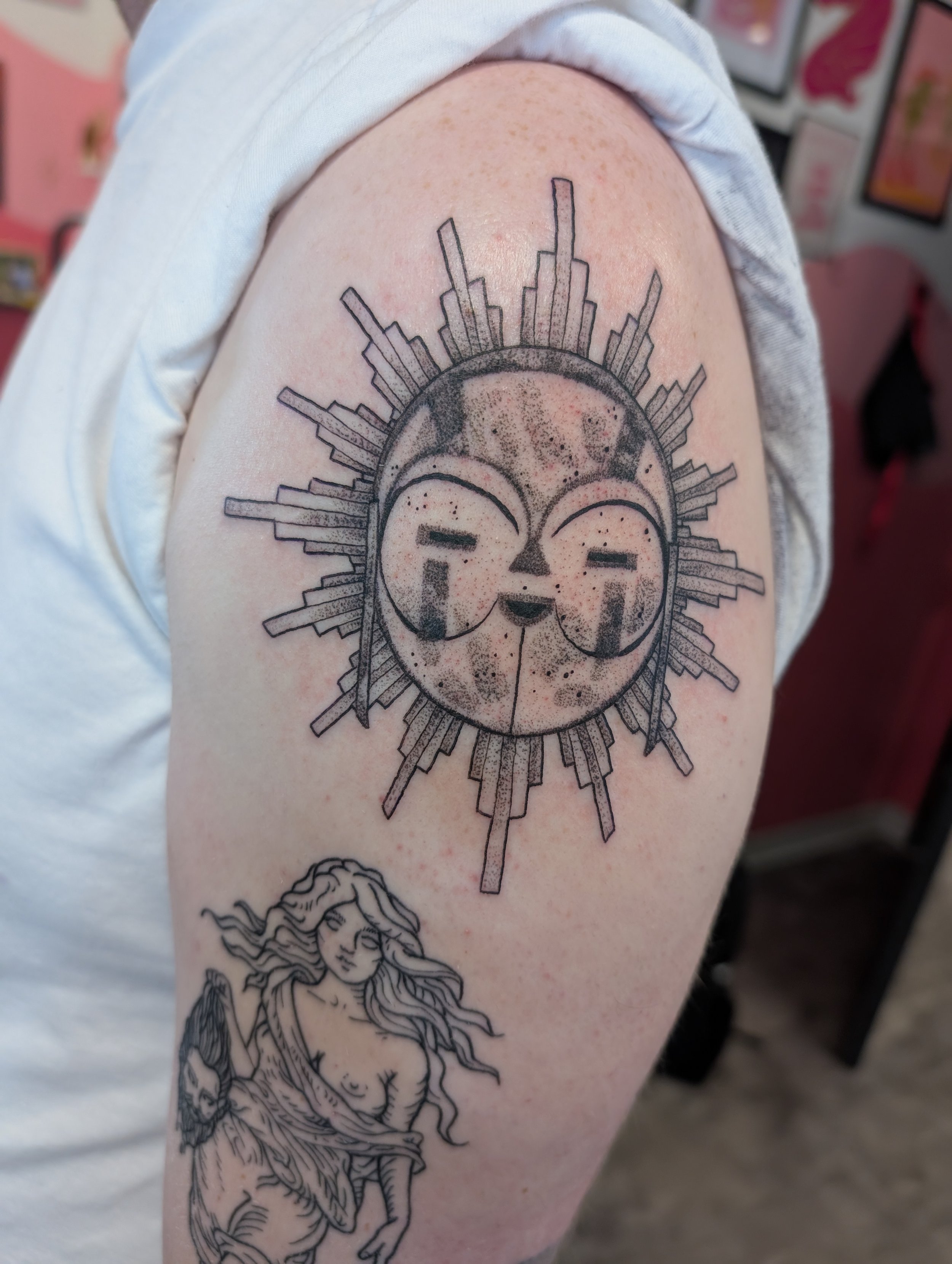 Tattoo of a skull with city skyline and a sunburst design on person's upper arm, alongside a depiction of a woman with long flowing hair.