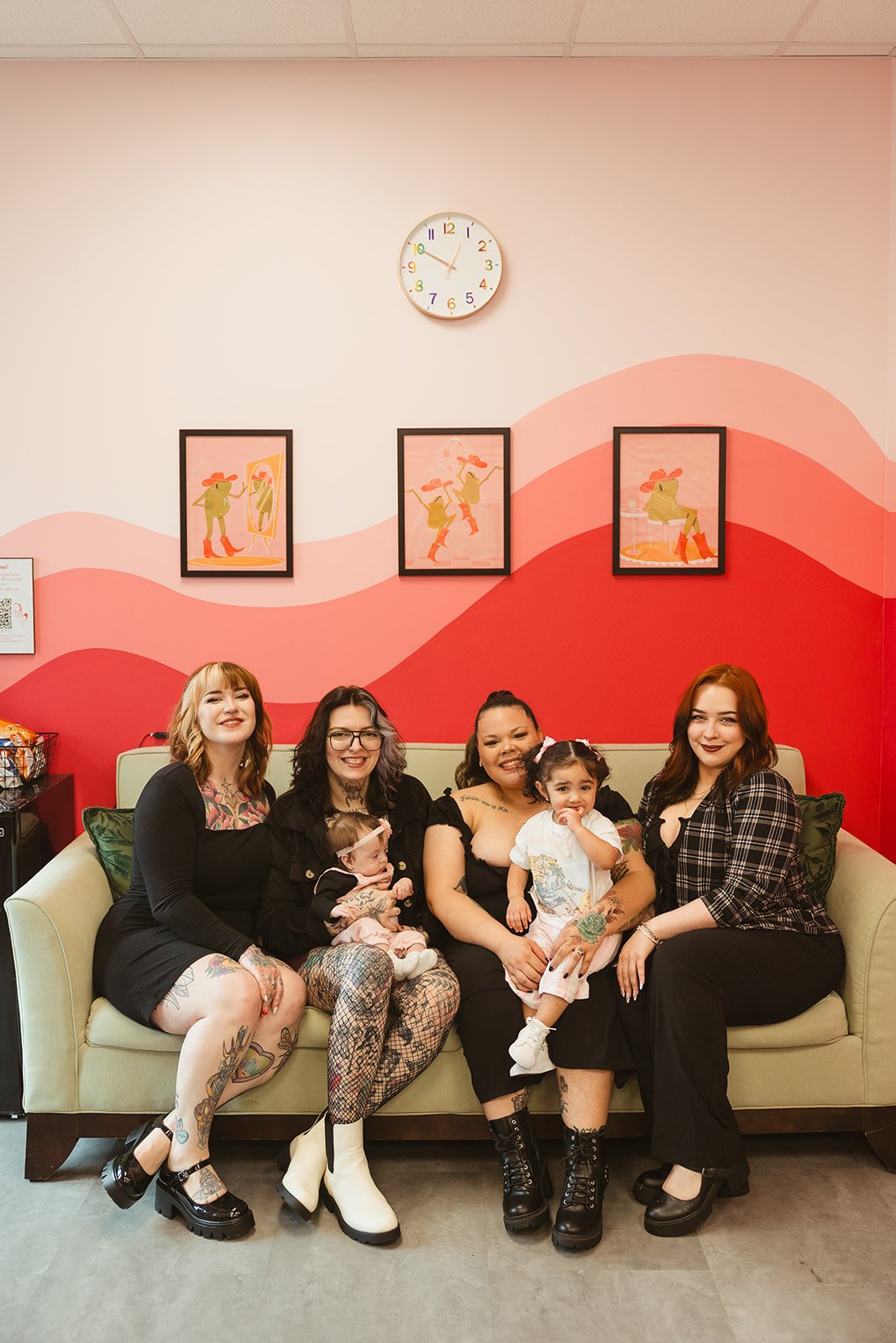 Group of beautiful women and their children sitting on a sofa in a brightly decorated room with pink and red wall art and framed pictures, smiling for the photo.