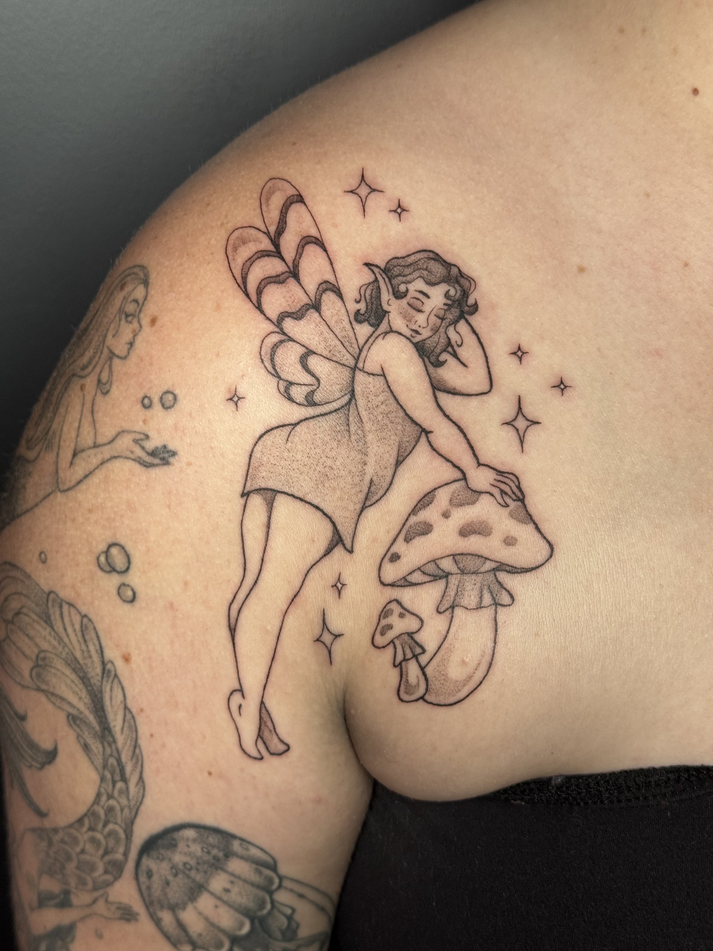 Tattoo of a fairy with butterfly wings perched on a mushroom, stars and other tattoo elements visible on the skin.