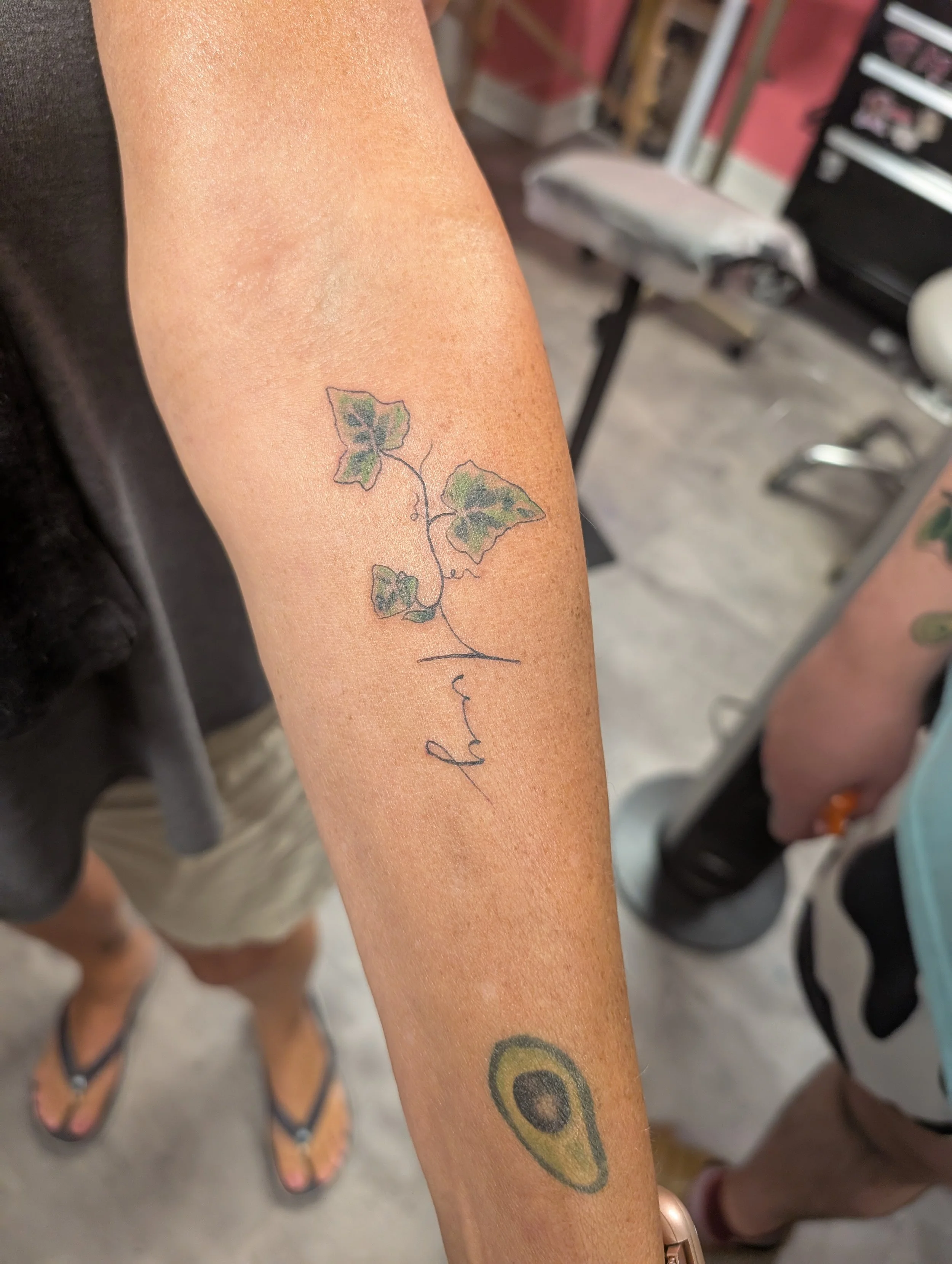 Close-up of a person's arm with tattoos. One tattoo of an avocado and another of ivy leaves. The person is wearing flip-flops and standing in what appears to be a tattoo studio or similar setting.