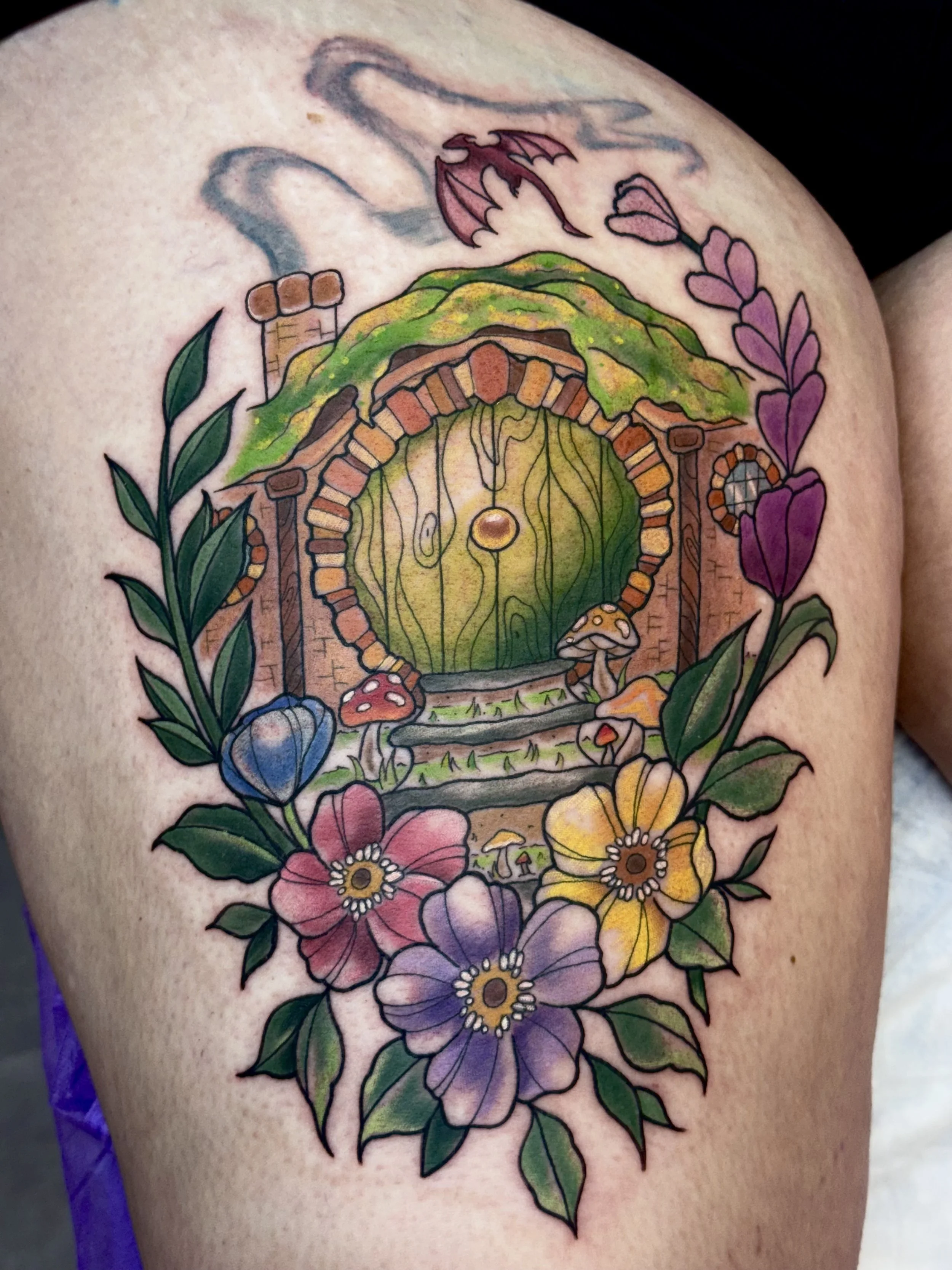 Colorful tattoo of a hobbit door surrounded by flowers, leaves, a dragon, and mushrooms on someone's leg.