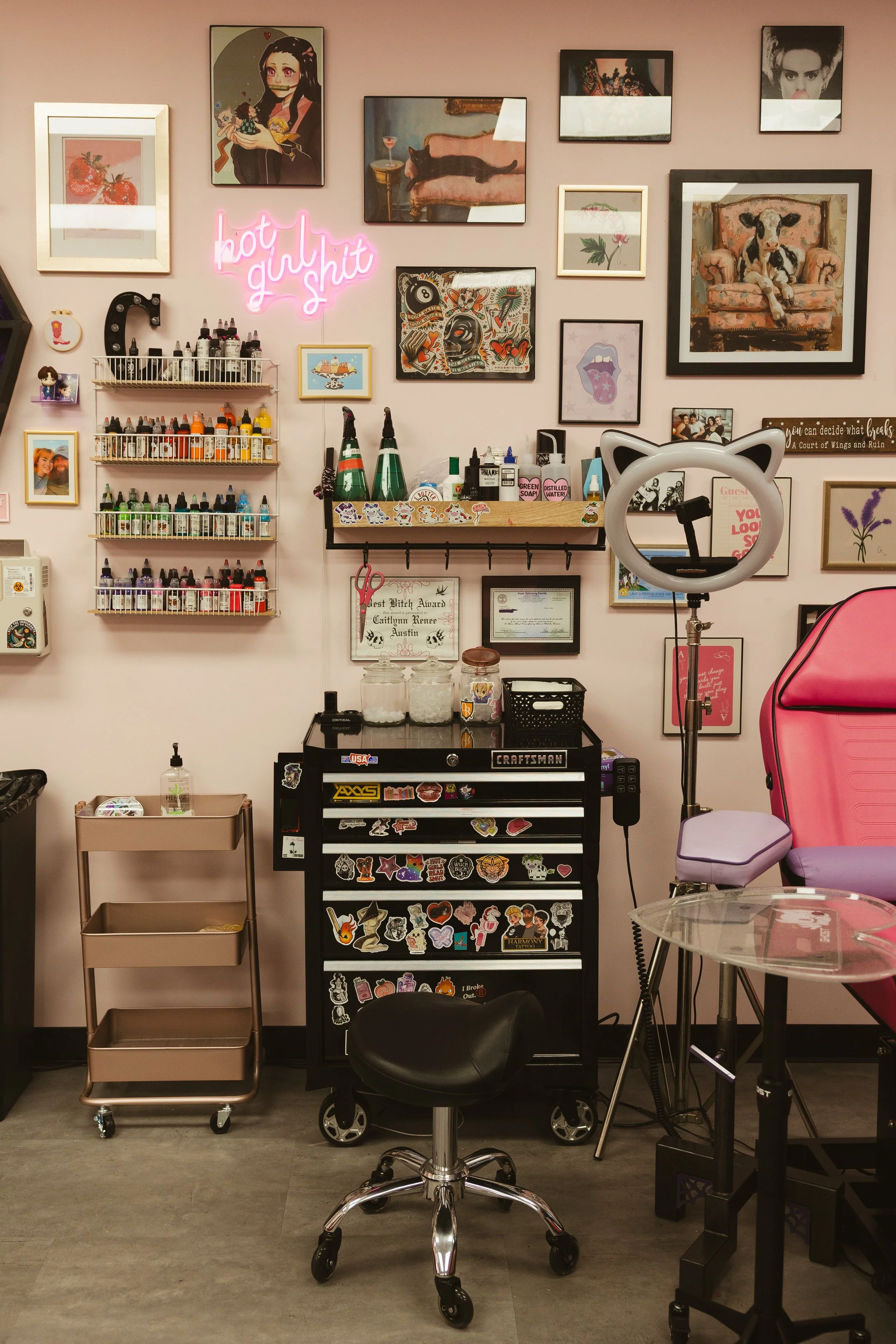 Wall with various framed pictures, neon sign reading 'hot girl shit', shelves with ink bottles, salon chairs, and equipment in a tattoo shop.