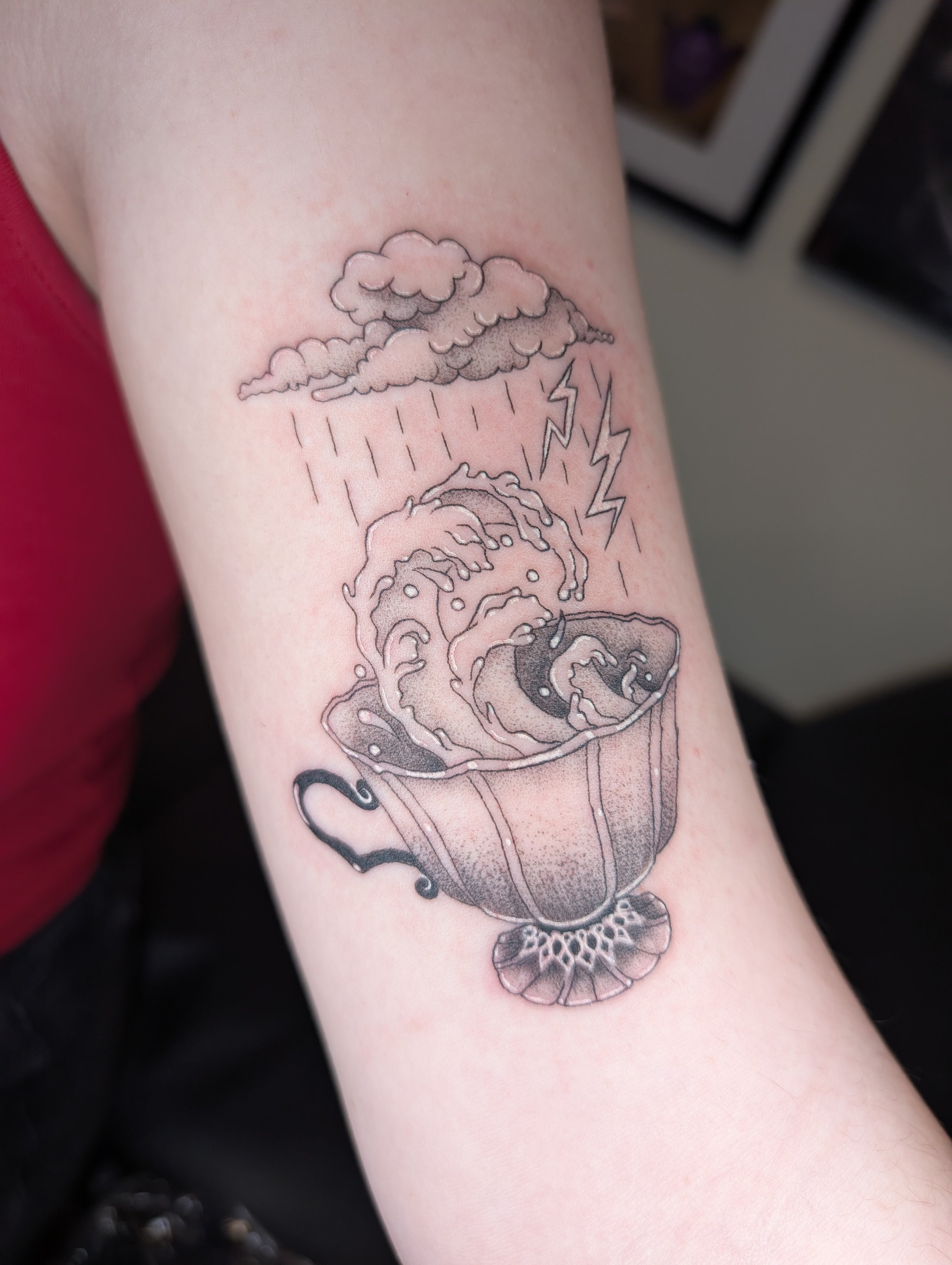 A black and gray tattoo of a steaming cup of coffee with a cloud of steam and rain, and lightning bolts, on a person's arm.