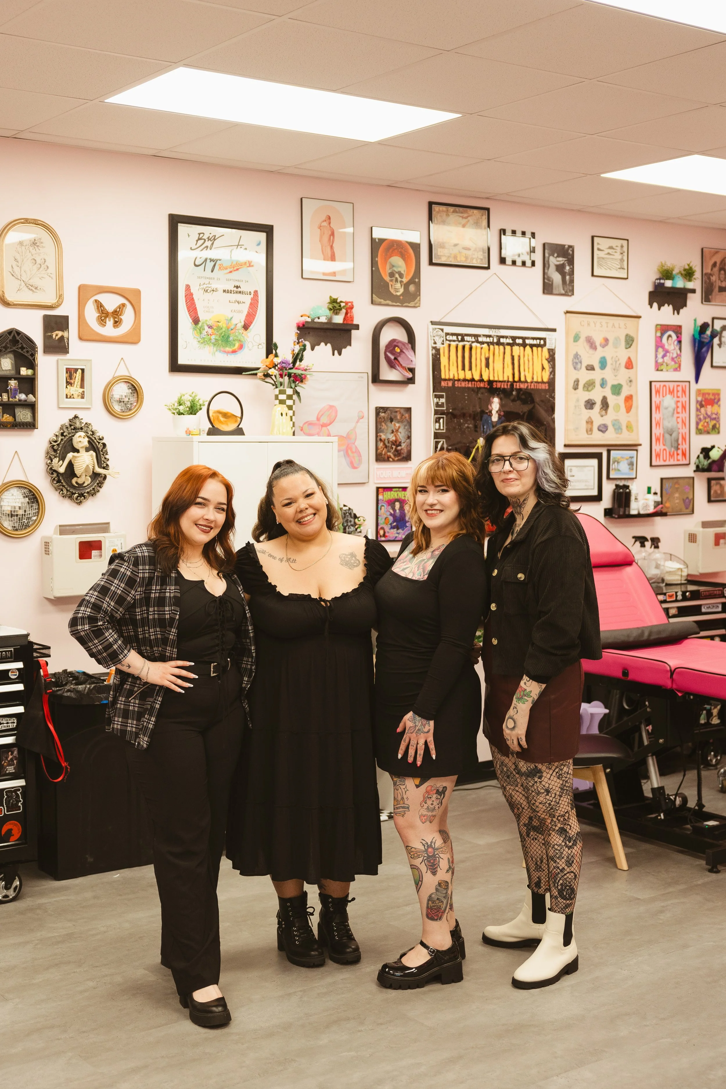 Four women standing together in an art-filled room, smiling at the camera. They are dressed in alternative fashion with tattoos and accessories, surrounded by various framed art and decorative objects on the walls.