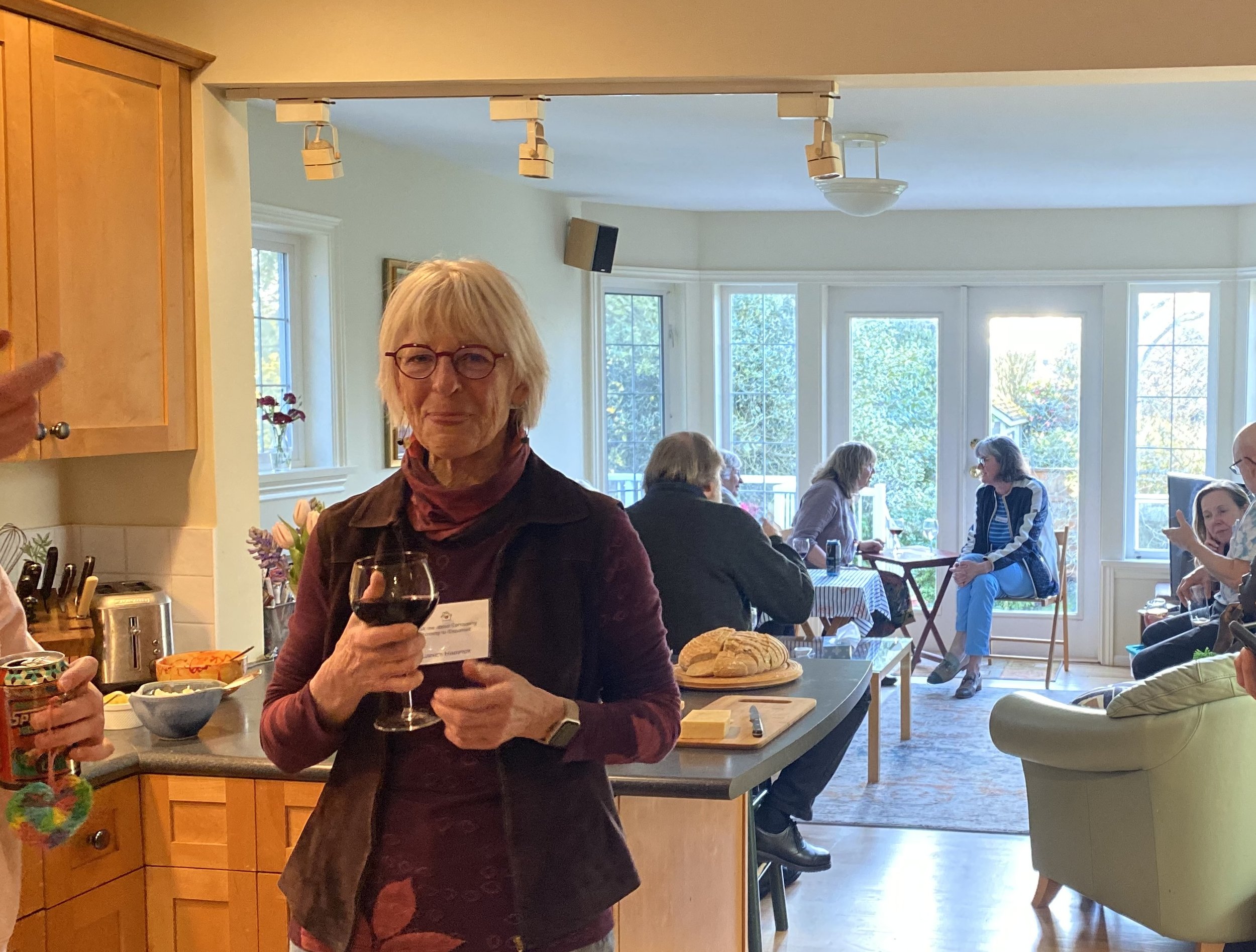 One of the founding members of Esquimalt Village Cohousing stands in her kitchen, smiling at you while holding a glass of red wine. In the background, people chat in an airy and bright living room over glasses of wine and appetizers.