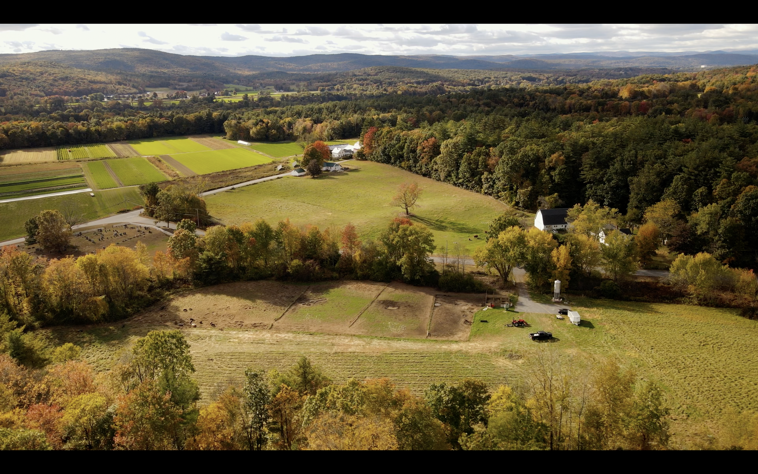 Aerial view of a rural landscape with farmland, wooded areas, and a few buildings, captured on a partly cloudy day in autumn.