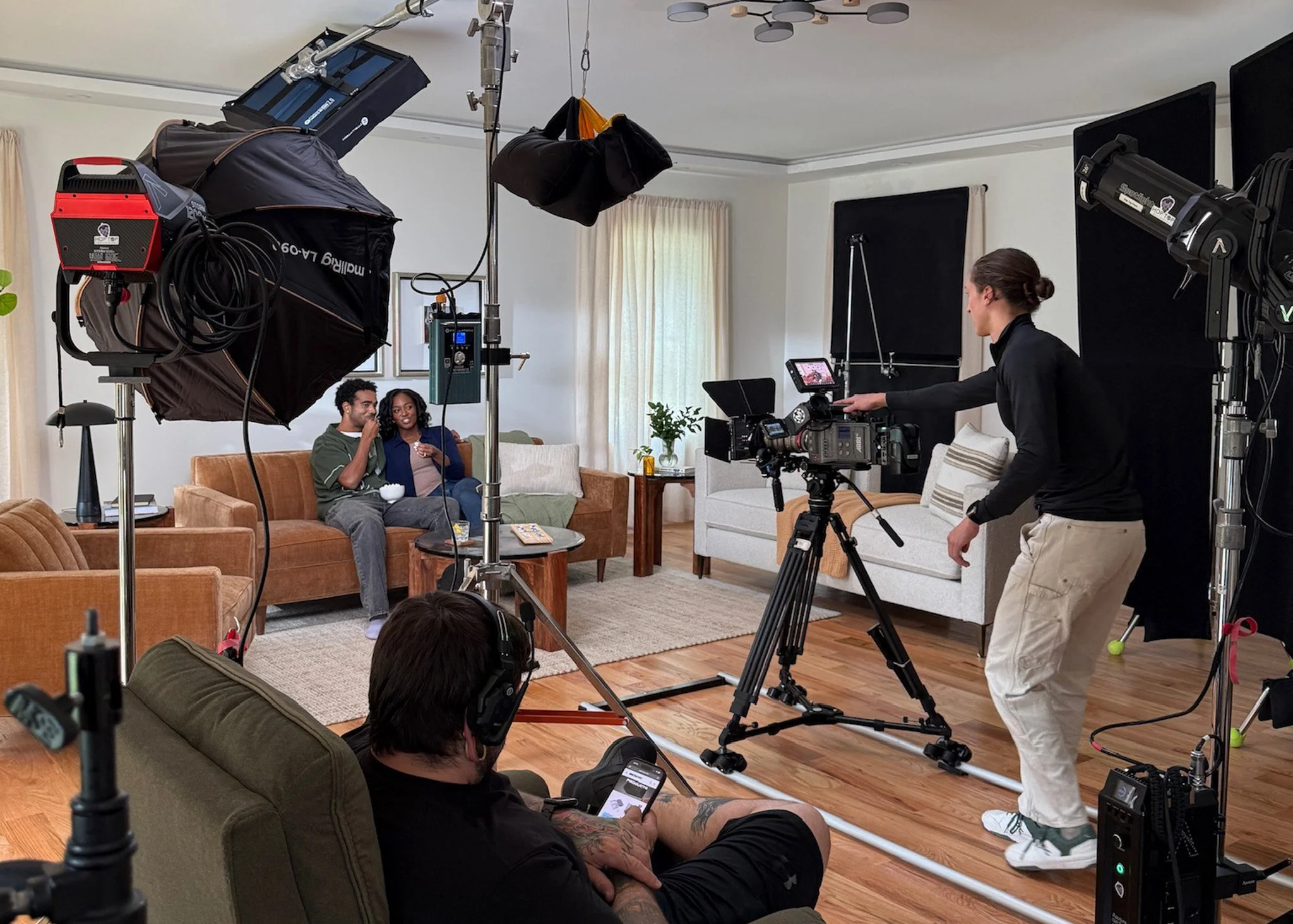 A film crew is shooting a scene in a living room with two people on a couch, while a woman adjusts camera equipment. The room has a white wall, beige curtains, and wooden flooring.