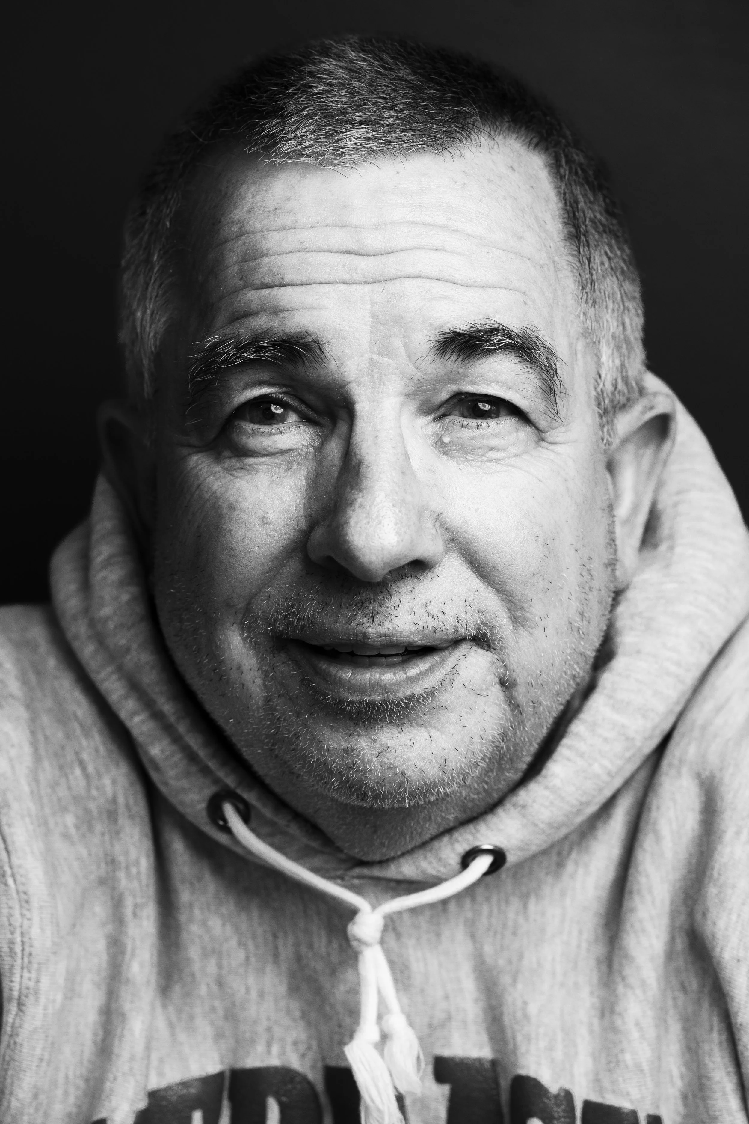Close-up black and white photo of a smiling man with short hair, wearing a hoodie.