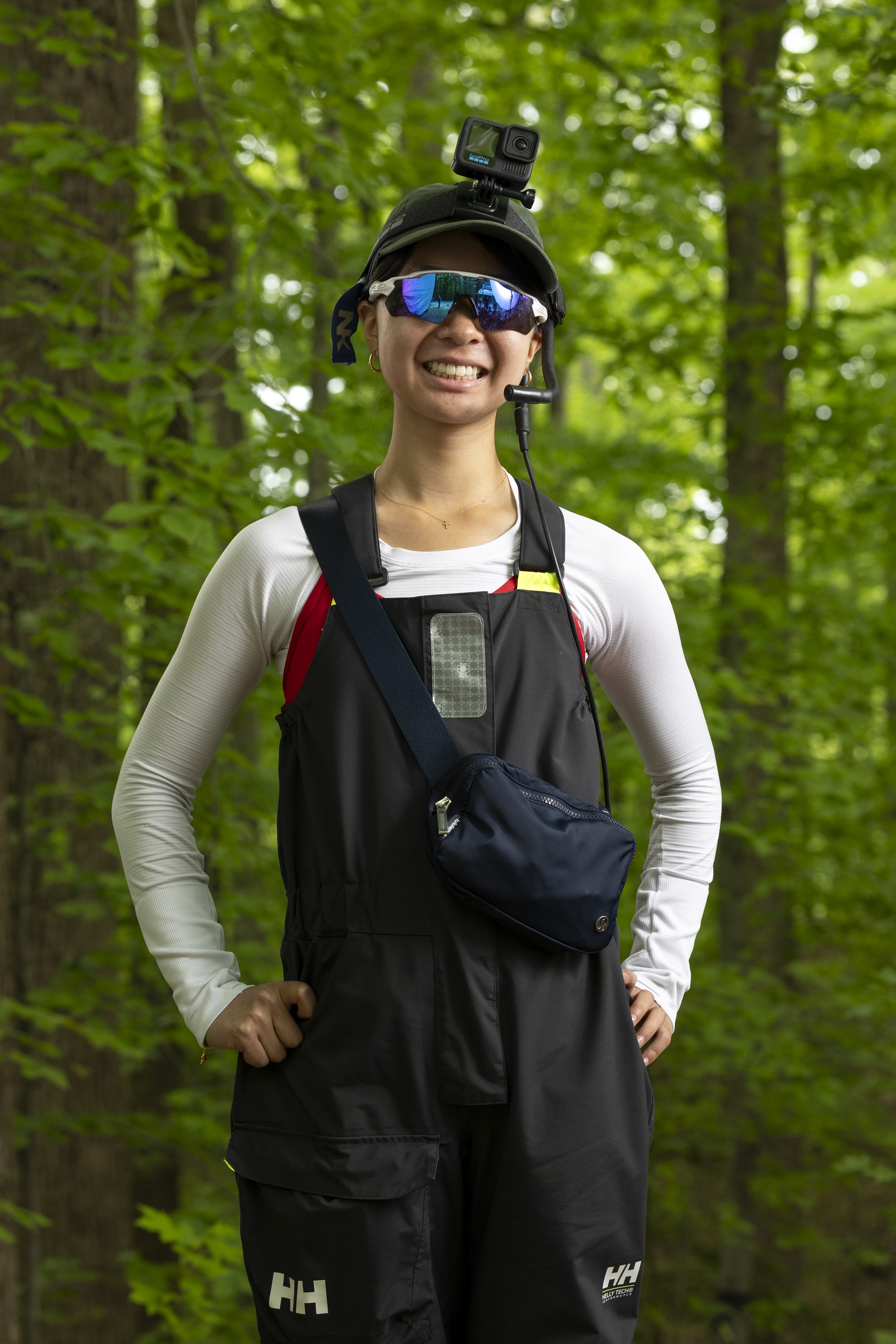 A woman in outdoor hiking gear standing in a green forest, wearing sunglasses, a cap with a GoPro camera mounted on top, a long-sleeve white shirt, a black vest with a small pouch, and black pants with white lettering, smiling at the camera.