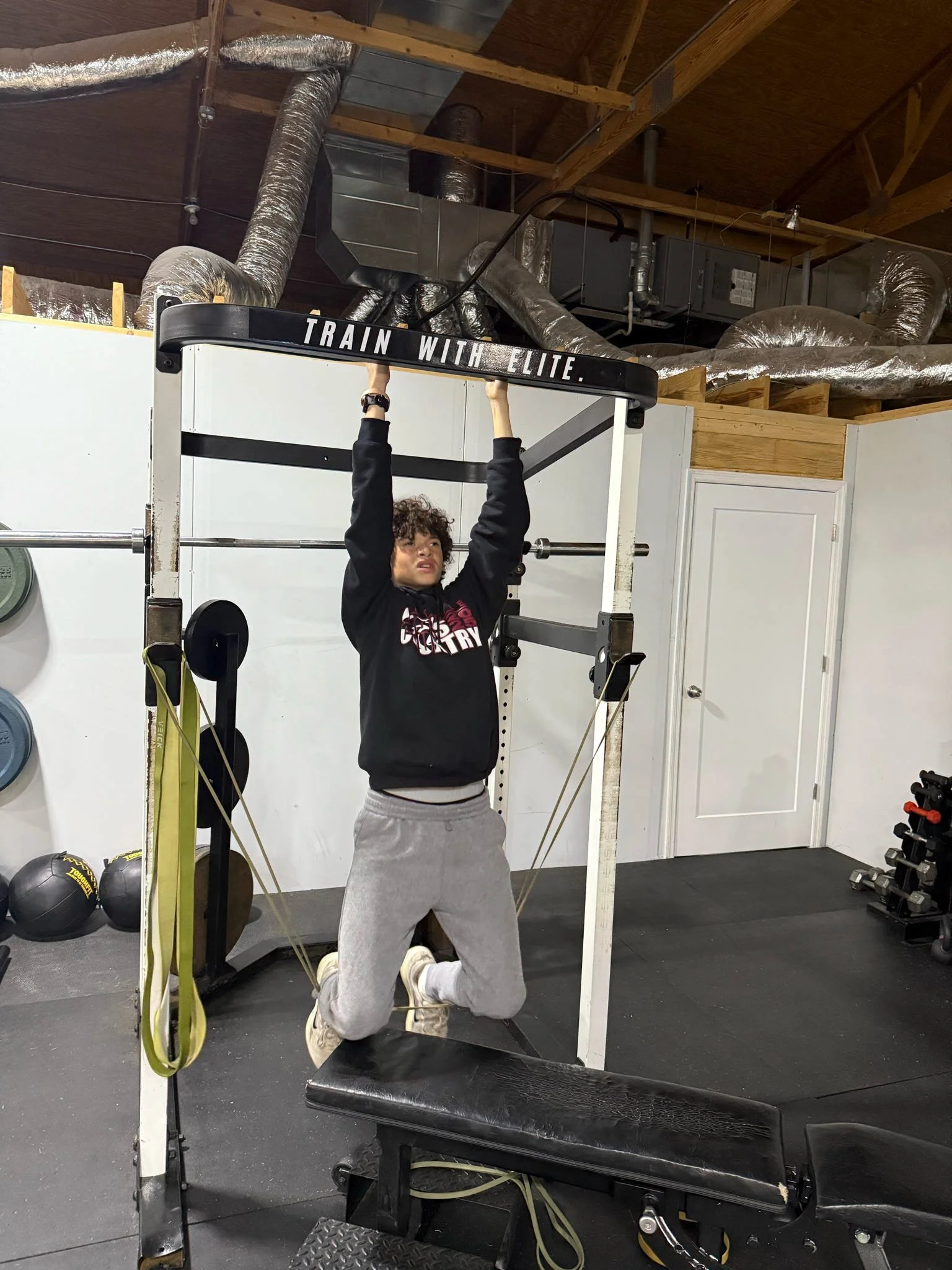 A person kneeling on one knee in a gym, holding onto a pull-up bar that has a sign reading 'Train with Elite.' The gym has weights, resistance bands, medicine balls, and gym equipment visible. The ceiling shows exposed ductwork and unfinished wood be