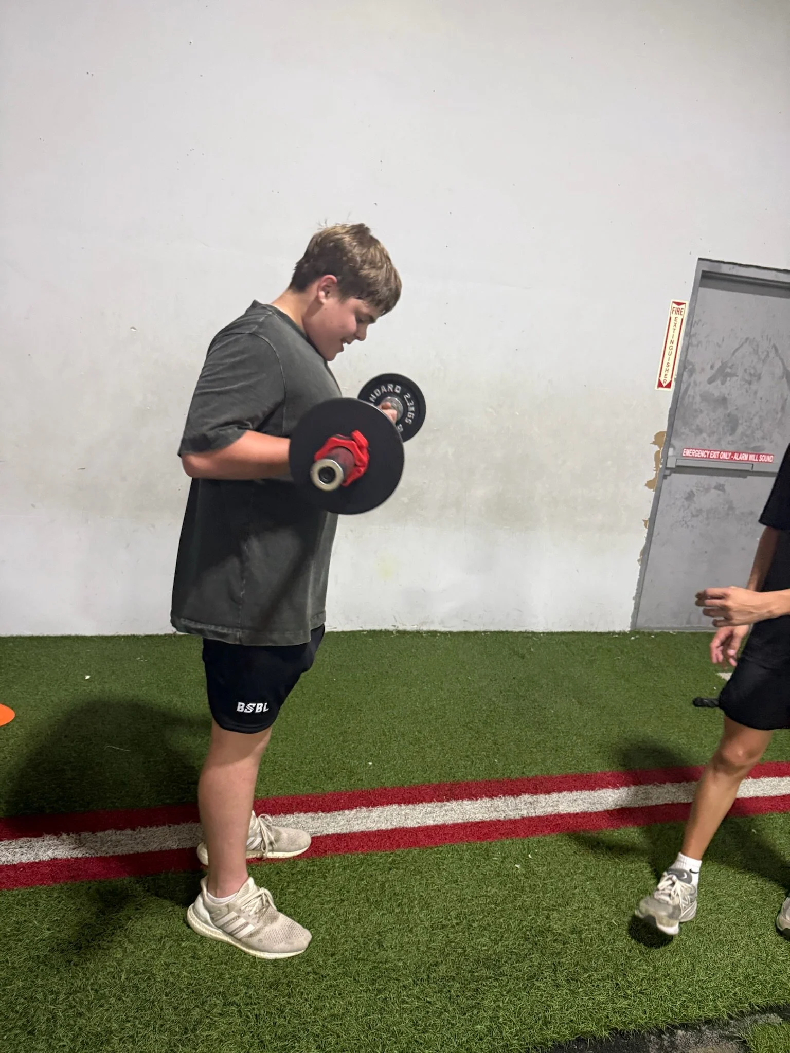 A young boy lifting a dumbbell while a woman observes in an indoor athletic facility with artificial turf.