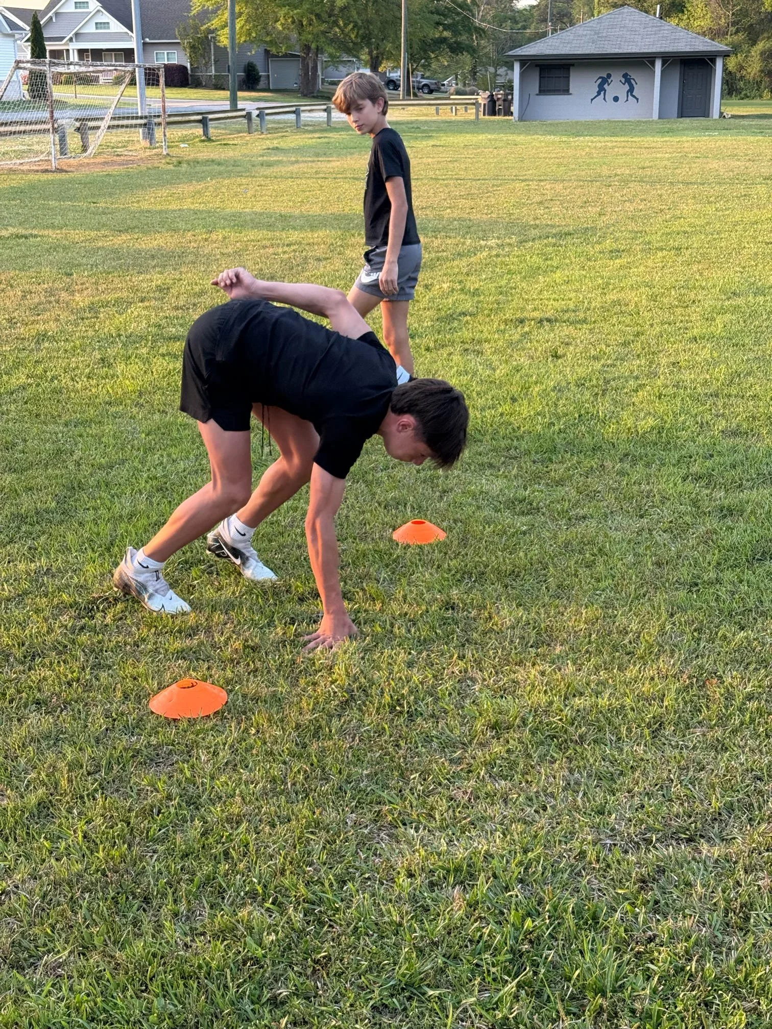 Two boys practicing athletic drills on a grassy field, one bending forward and the other standing behind, with orange cones set up for training, in a park or schoolyard setting.