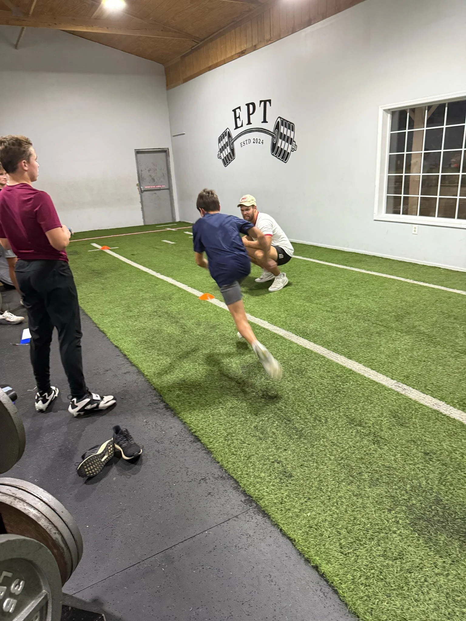 Group of young men at indoor gym, one is squatting with a trainer or coach giving instructions, and others are watching. The gym has a wall with a logo of a barbell and the text 'EPT' and 'ESTD 2024'. The floor has green turf and black rubber matting