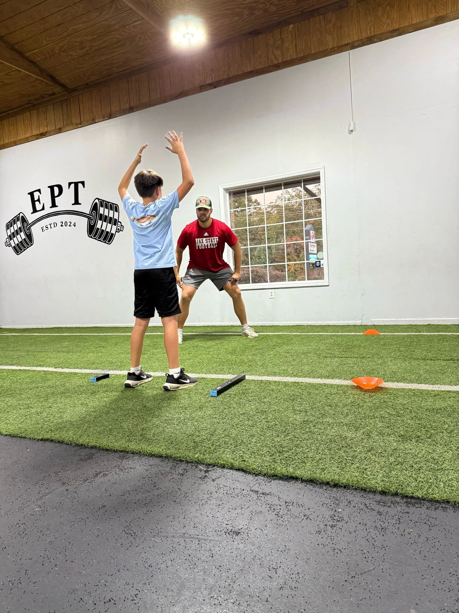 A young boy and an adult male are inside a gym. The boy is facing away with his arms raised, and the adult is standing in front of him, crouched with hands on his knees, preparing for a football practice or drill. The goal wall on the left has a logo