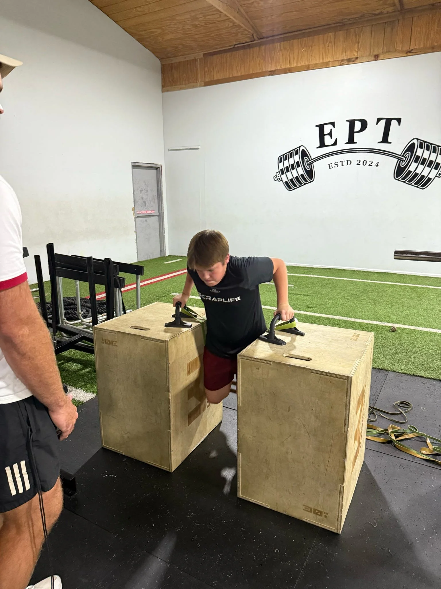 A young boy is doing push-ups with his hands on two wooden plyometric boxes in a gym, with a logo on the wall reading 'EPT ESTD 2024' alongside a barbell graphic.