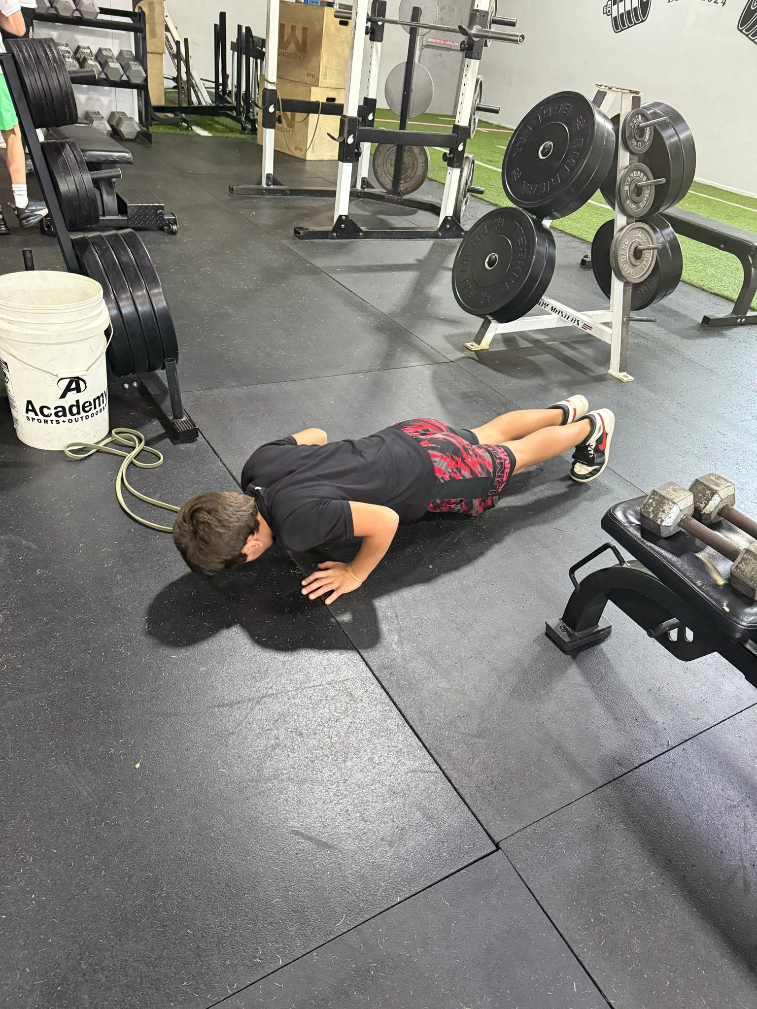 A person doing a push-up in a gym with various weightlifting equipment, dumbbells, and weight plates visible.