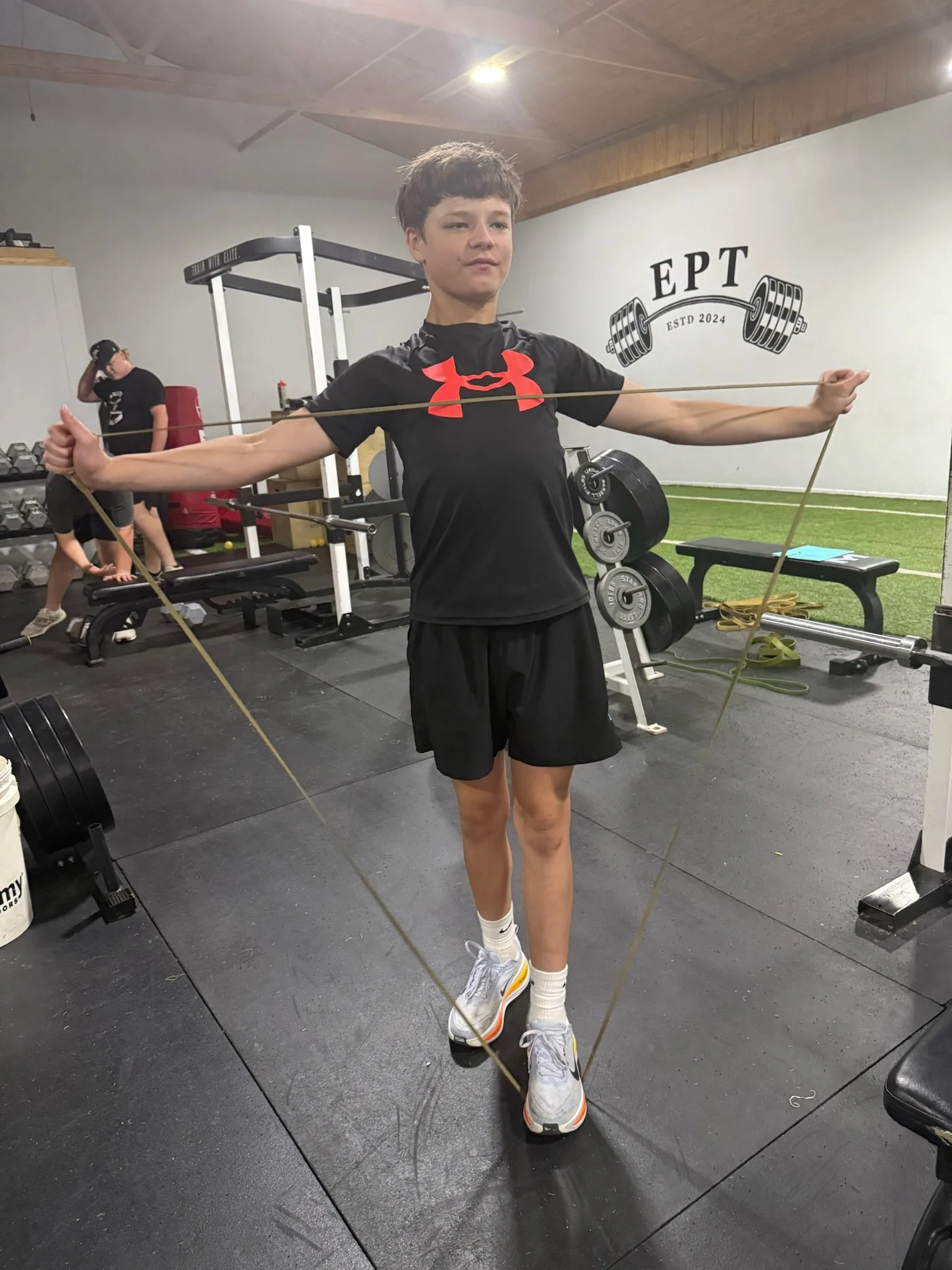 A young person with short hair wearing a black Under Armour shirt, black shorts, and white sneakers with orange accents is standing in a gym, holding a resistance band with both hands and pressing it outwards. There are weights, a workout bench, and 