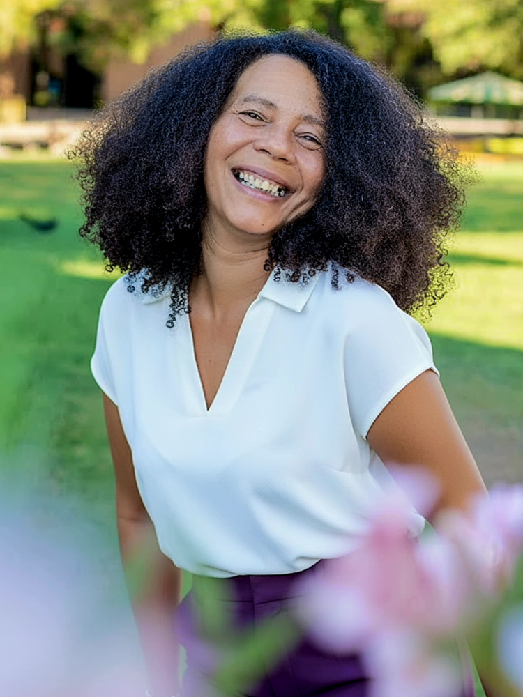 A woman with curly black hair and light brown skin, wearing a peach-colored top and layered necklaces, is resting her head on a large piece of driftwood or tree branch outdoors. She has a gentle smile and is looking directly at the camera.