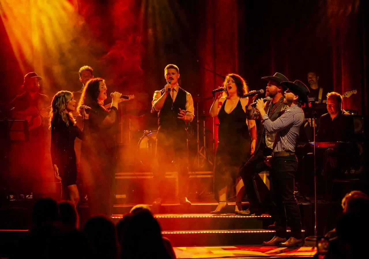 A group of musicians and singers perform on stage with dramatic red and orange lighting. They are surrounded by instruments and a live band, creating an engaging concert atmosphere.