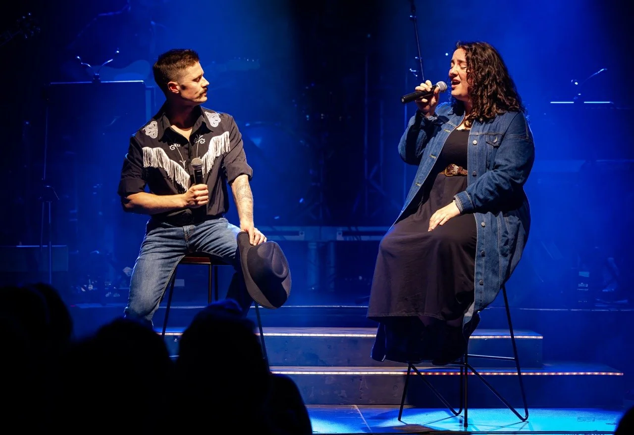 Two performers on stage with microphones, one holding a hat, under blue stage lighting.