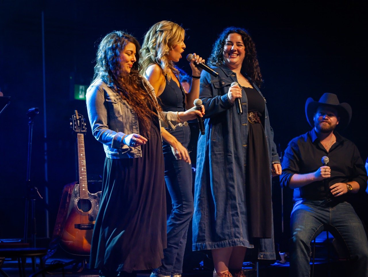 Group of people singing on stage with microphones, wearing casual clothing and denim. Acoustic guitar and music stand in the background.