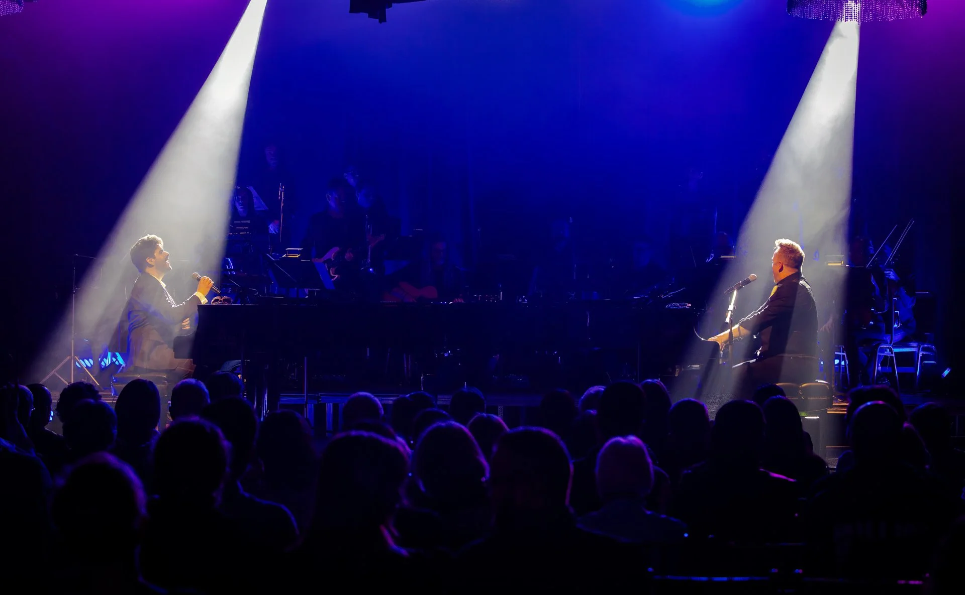 Two musicians performing on stage, each playing a piano and facing each other, illuminated by spotlight beams in a dark concert hall with an audience.