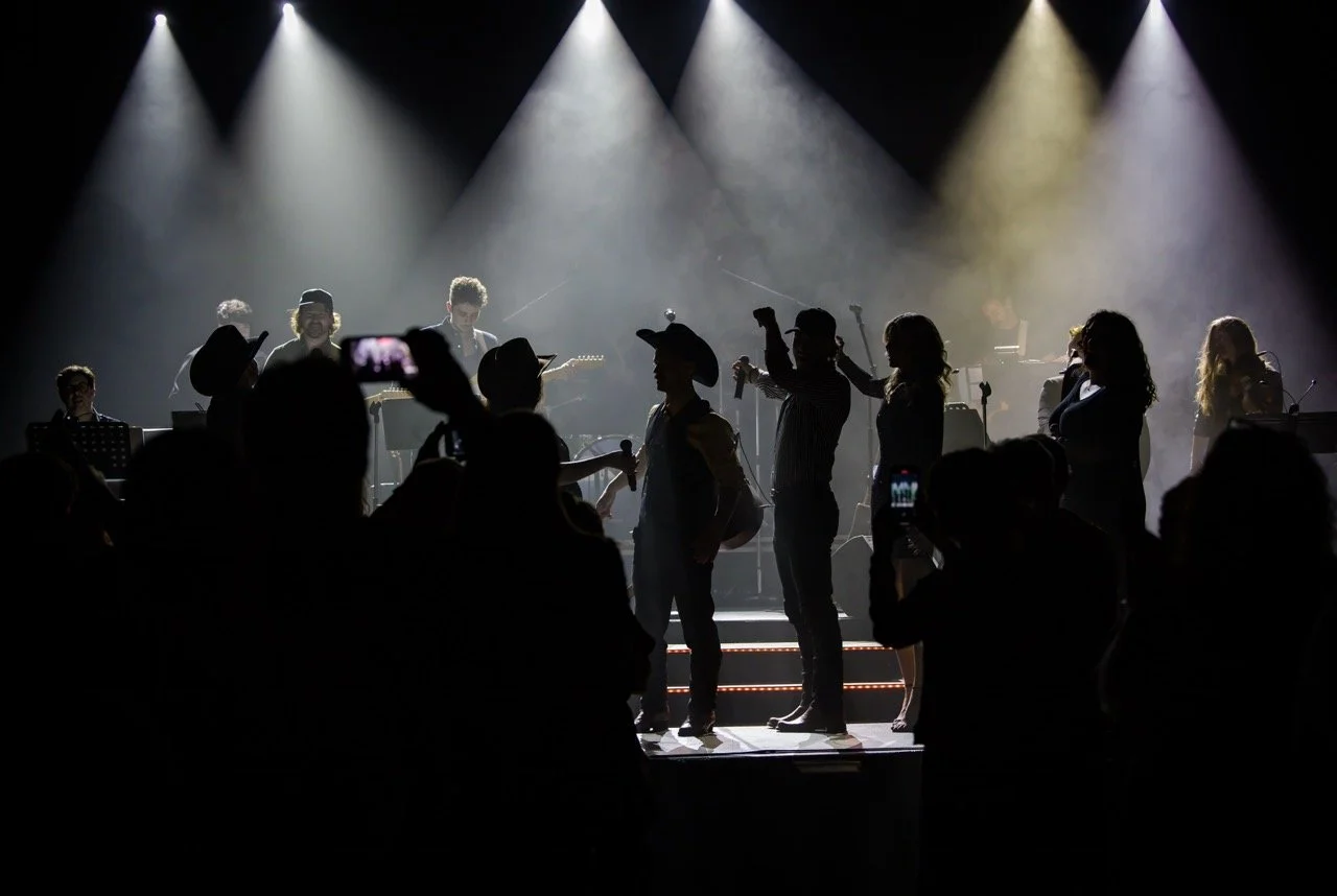 Silhouettes of performers on a dimly lit stage, with audience members capturing the scene using smartphones, and stage lights creating a dramatic effect.