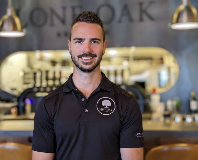 Smiling man in black polo shirt with "Lone Oak" logo, standing in a cafe or bar setting with pendant lights and bar counter in the background.