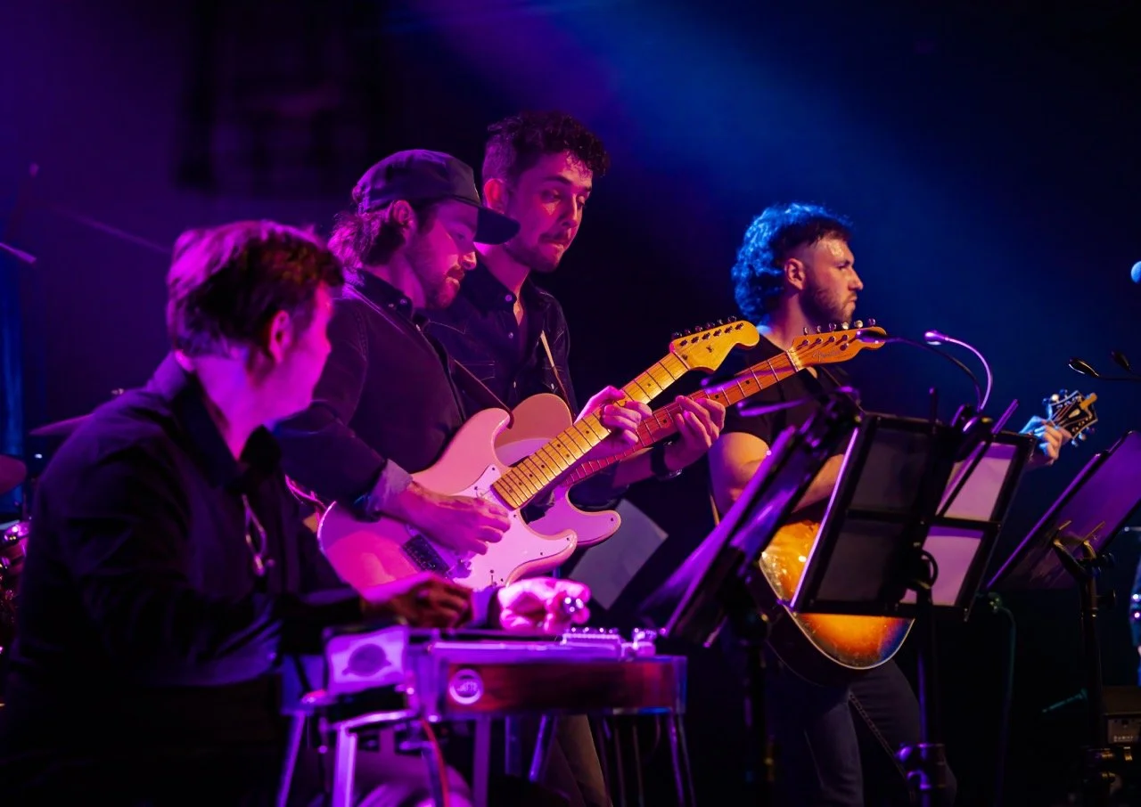 Four musicians performing on stage, playing electric and acoustic guitars, with music stands visible, under purple stage lighting.