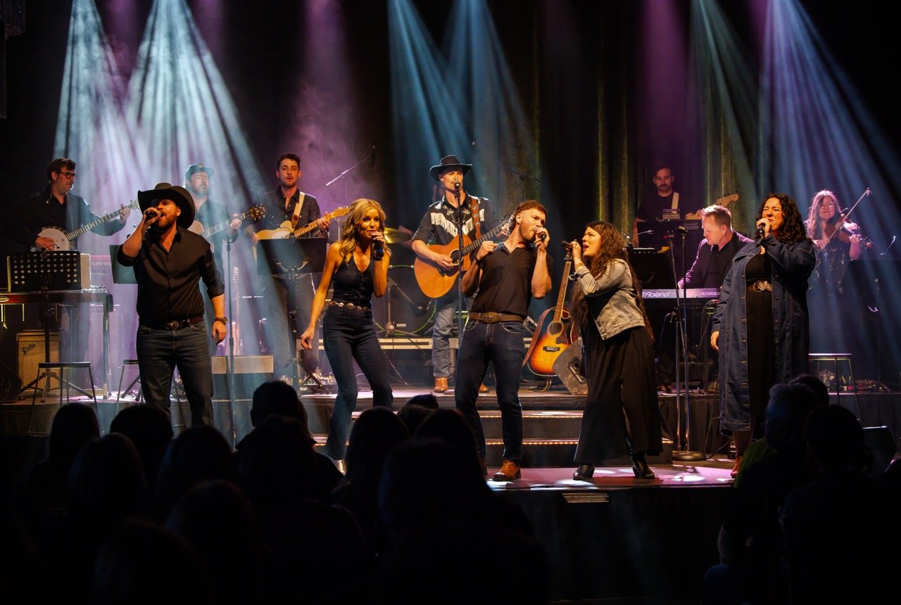 Band performing on stage with multiple singers and musicians playing instruments under colorful stage lights.