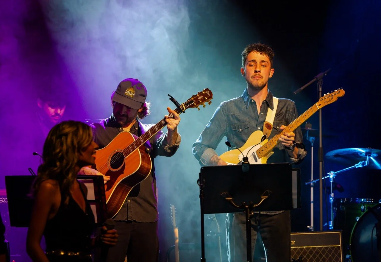Musicians performing on stage with guitars and microphone, colorful lighting, and mist effects.